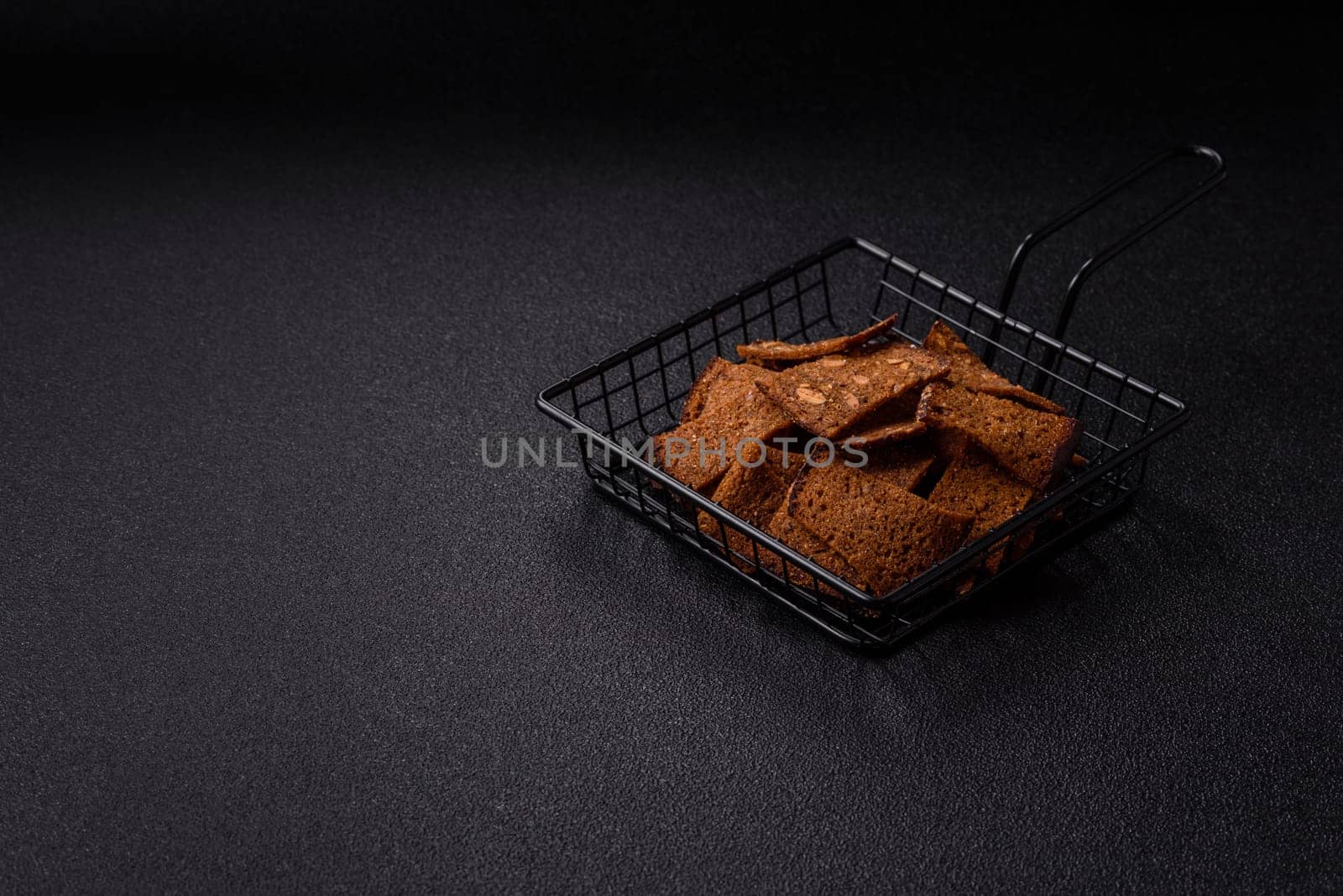 Crispy homemade brown bread chips with seeds and grains, salt, spices and herbs on a dark concrete background