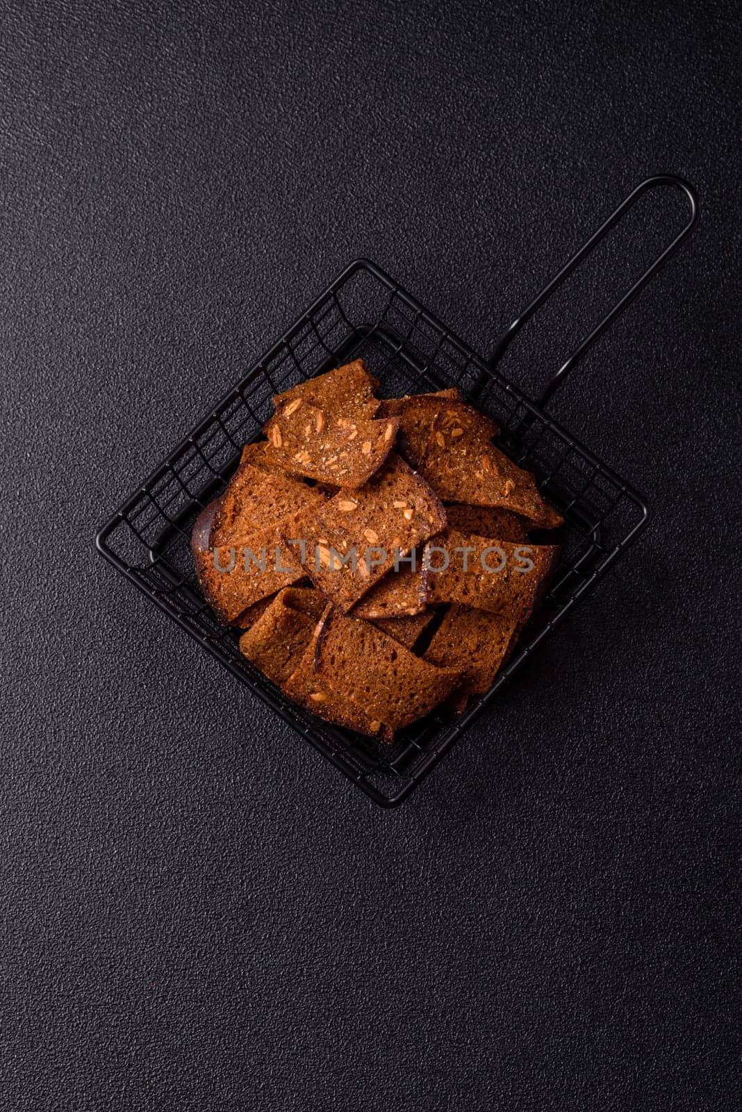 Crispy homemade brown bread chips with seeds and grains, salt, spices and herbs on a dark concrete background