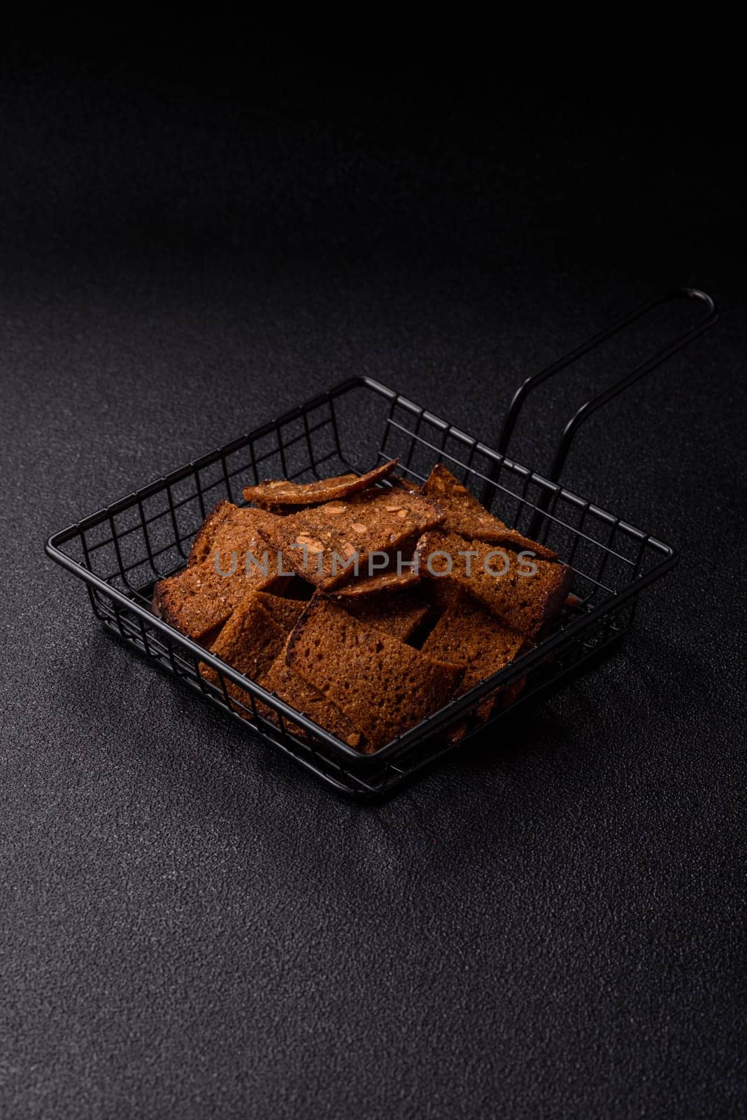 Crispy homemade brown bread chips with seeds and grains, salt, spices and herbs on a dark concrete background