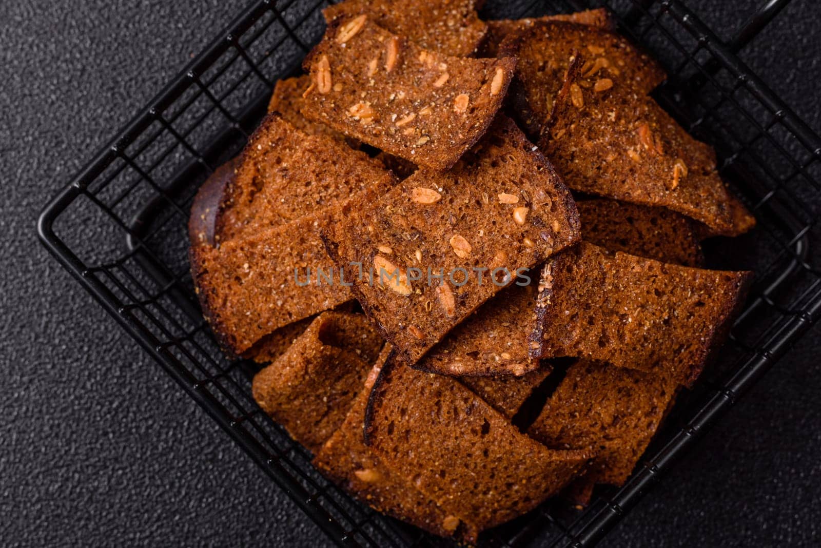 Crispy homemade brown bread chips with seeds and grains, salt, spices and herbs on a dark concrete background