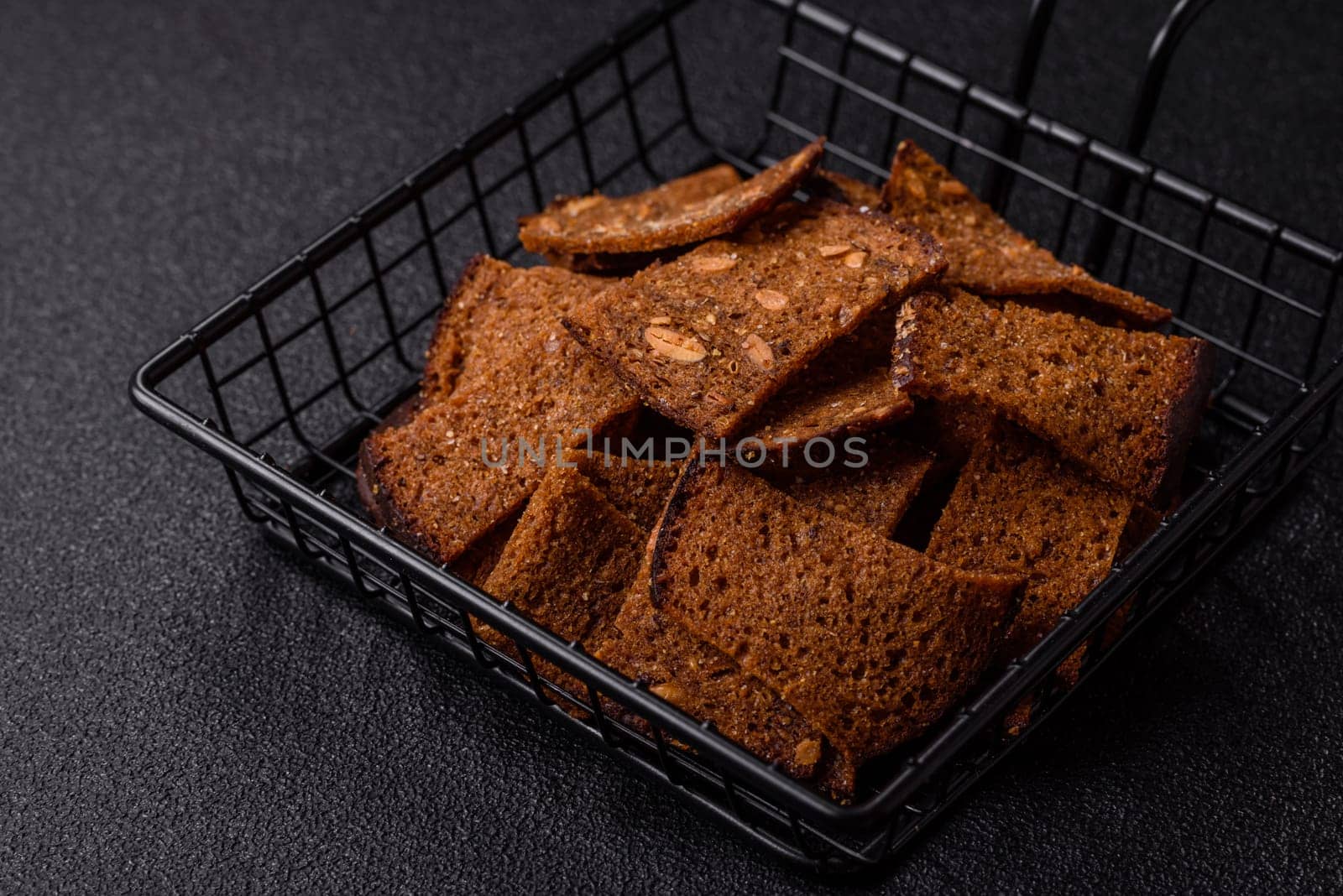 Crispy homemade brown bread chips with seeds and grains, salt, spices and herbs on a dark concrete background