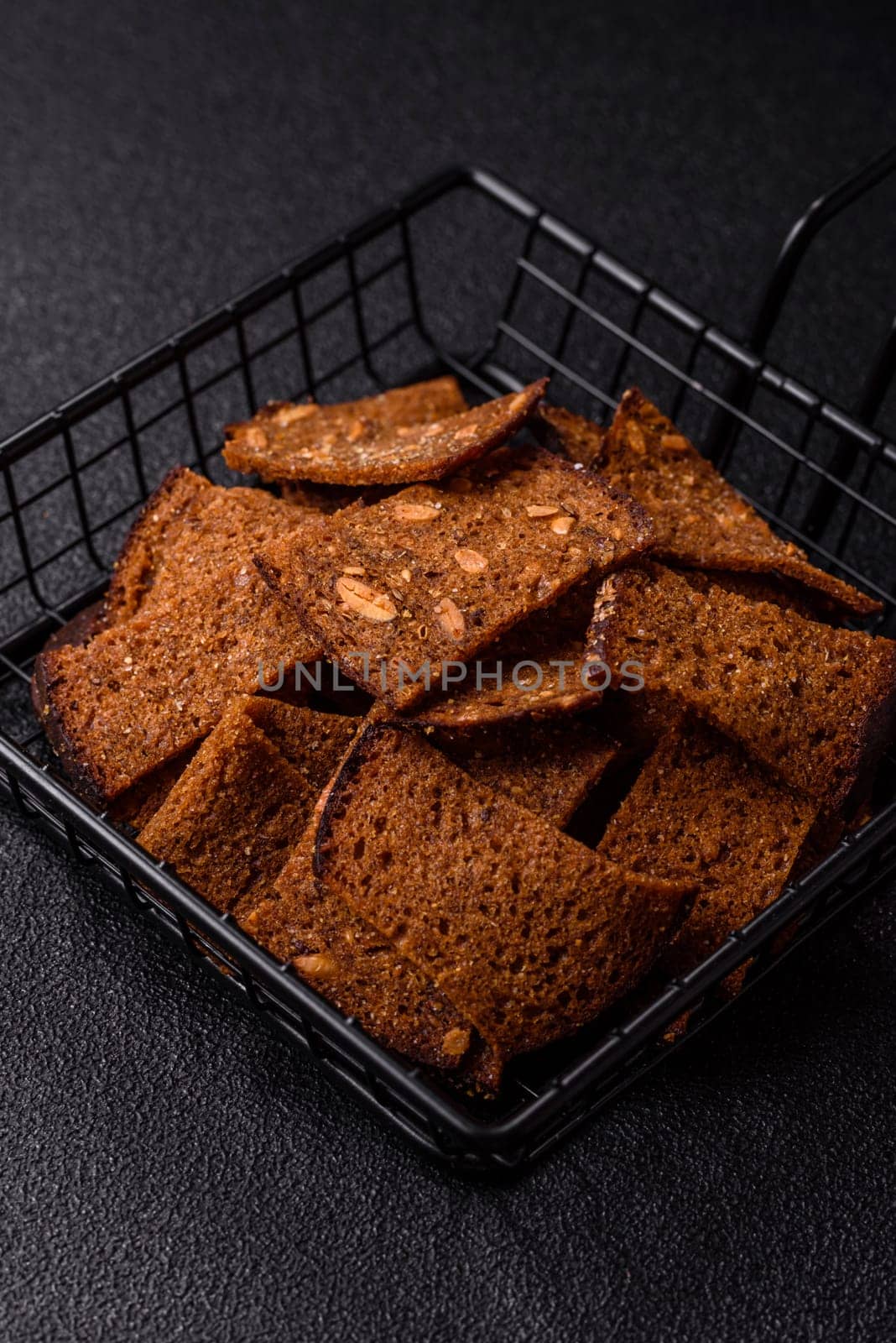 Crispy homemade brown bread chips with seeds and grains, salt, spices and herbs on a dark concrete background