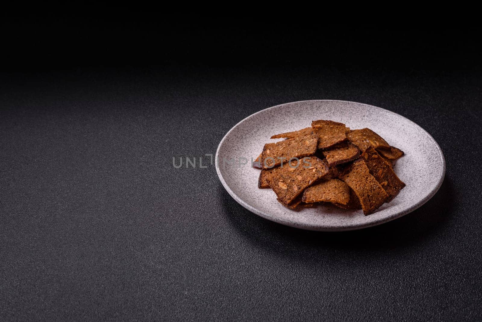 Crispy homemade brown bread chips with seeds and grains, salt, spices and herbs on a dark concrete background