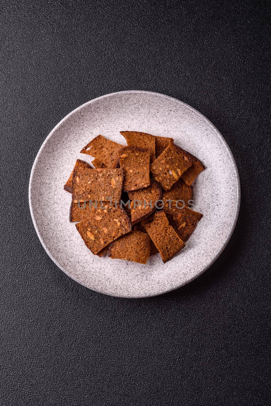 Crispy homemade brown bread chips with seeds and grains, salt, spices and herbs on a dark concrete background