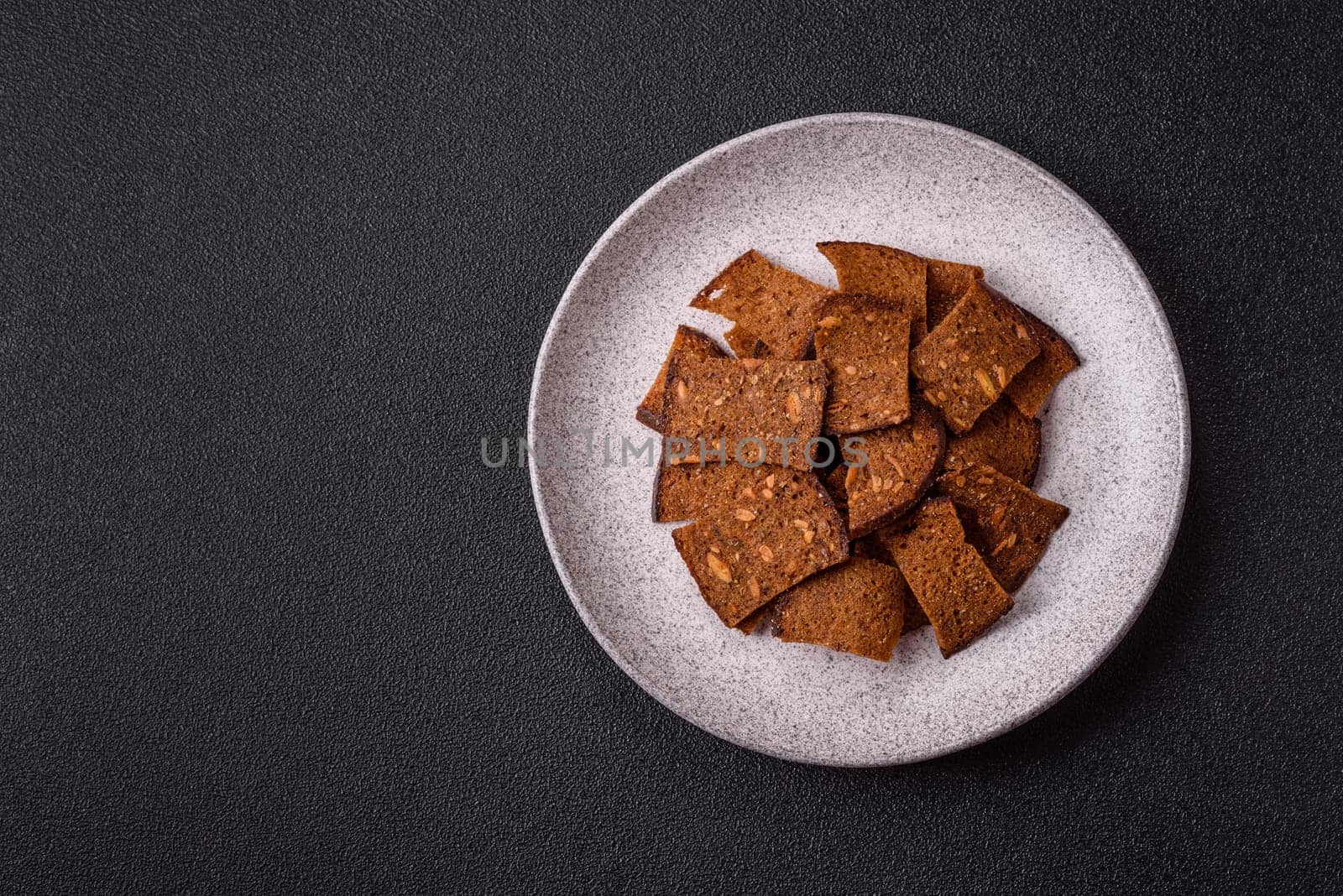Crispy homemade brown bread chips with seeds and grains, salt, spices and herbs on a dark concrete background