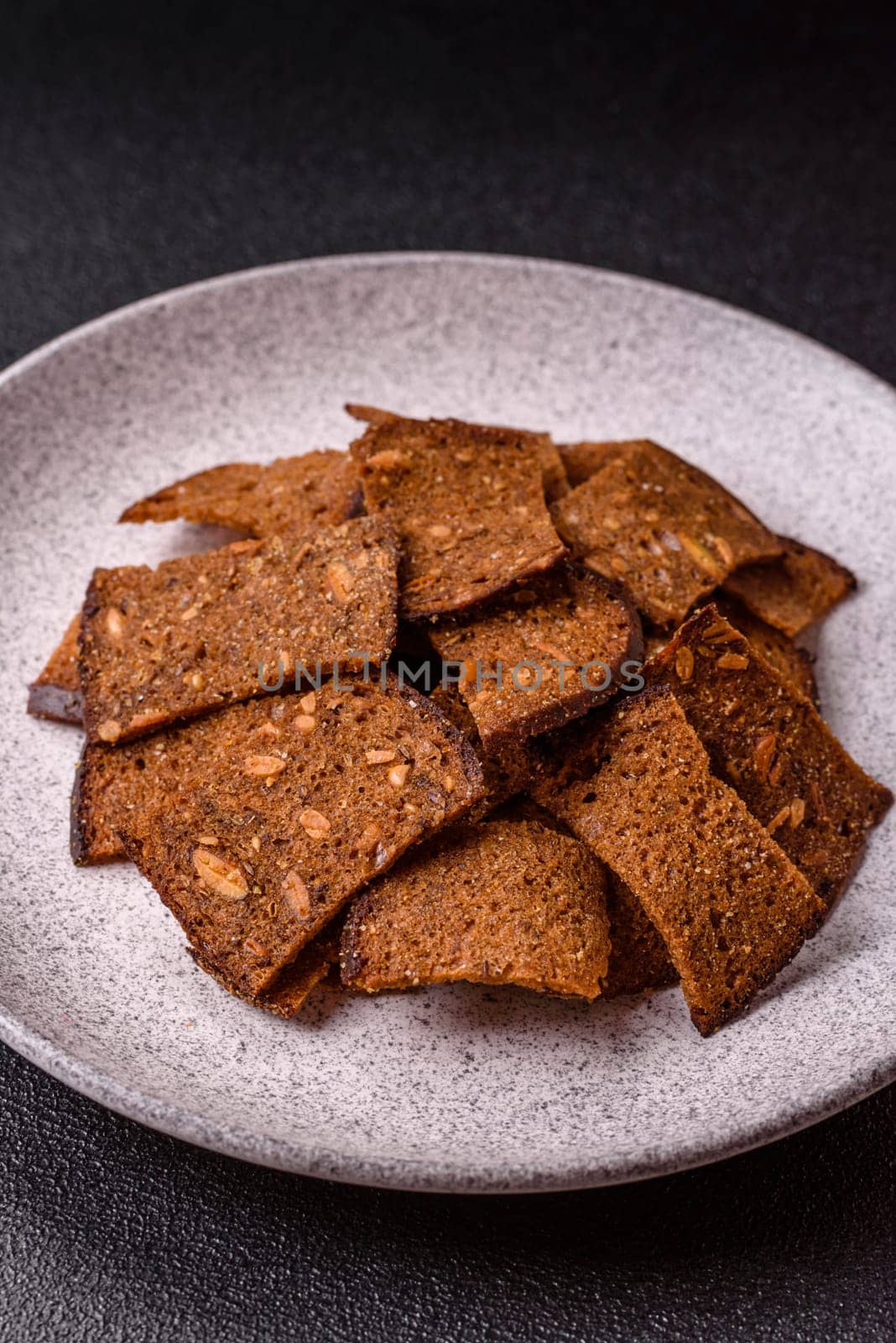 Crispy homemade brown bread chips with seeds and grains, salt, spices and herbs on a dark concrete background