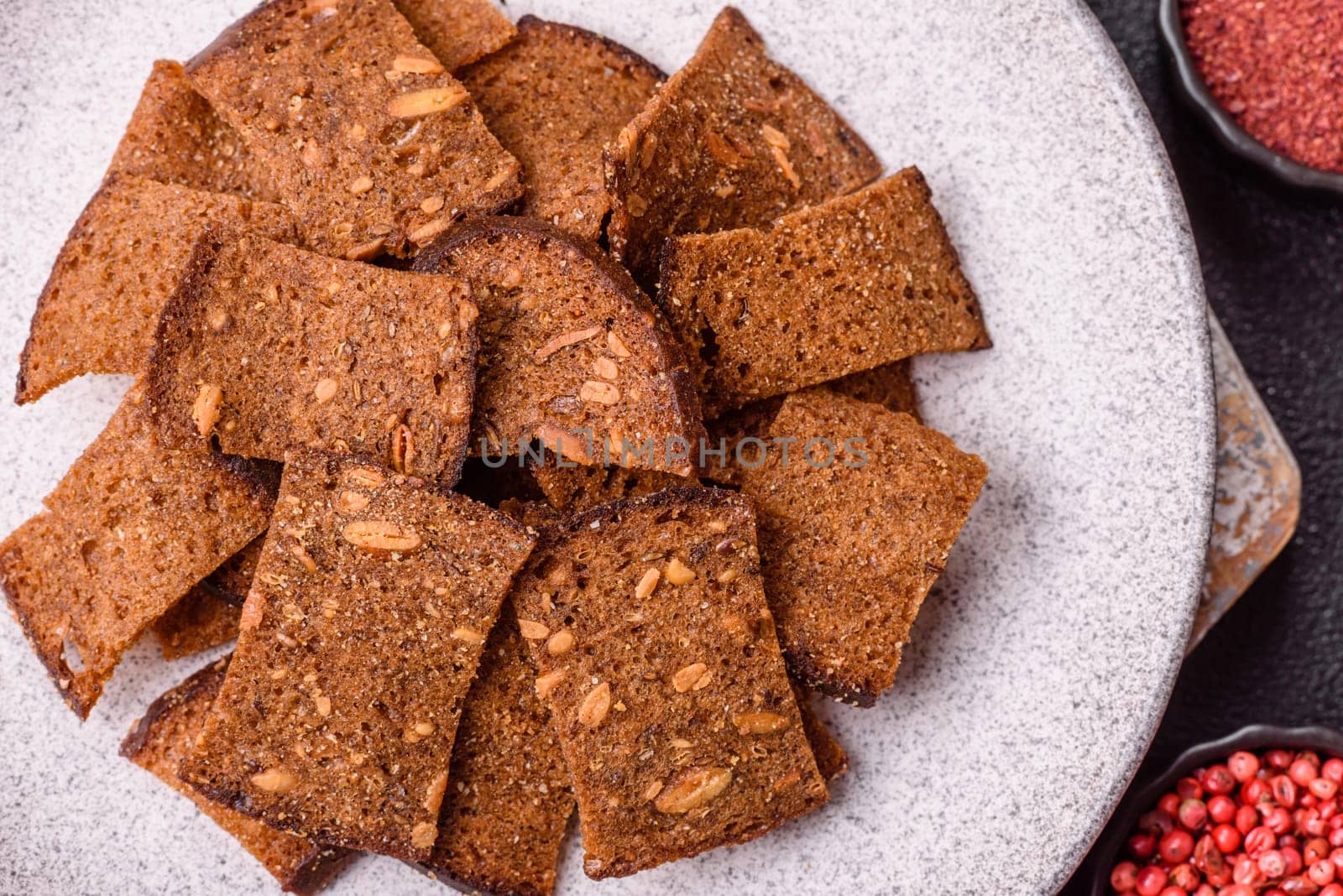 Crispy homemade brown bread chips with seeds and grains, salt, spices and herbs on a dark concrete background