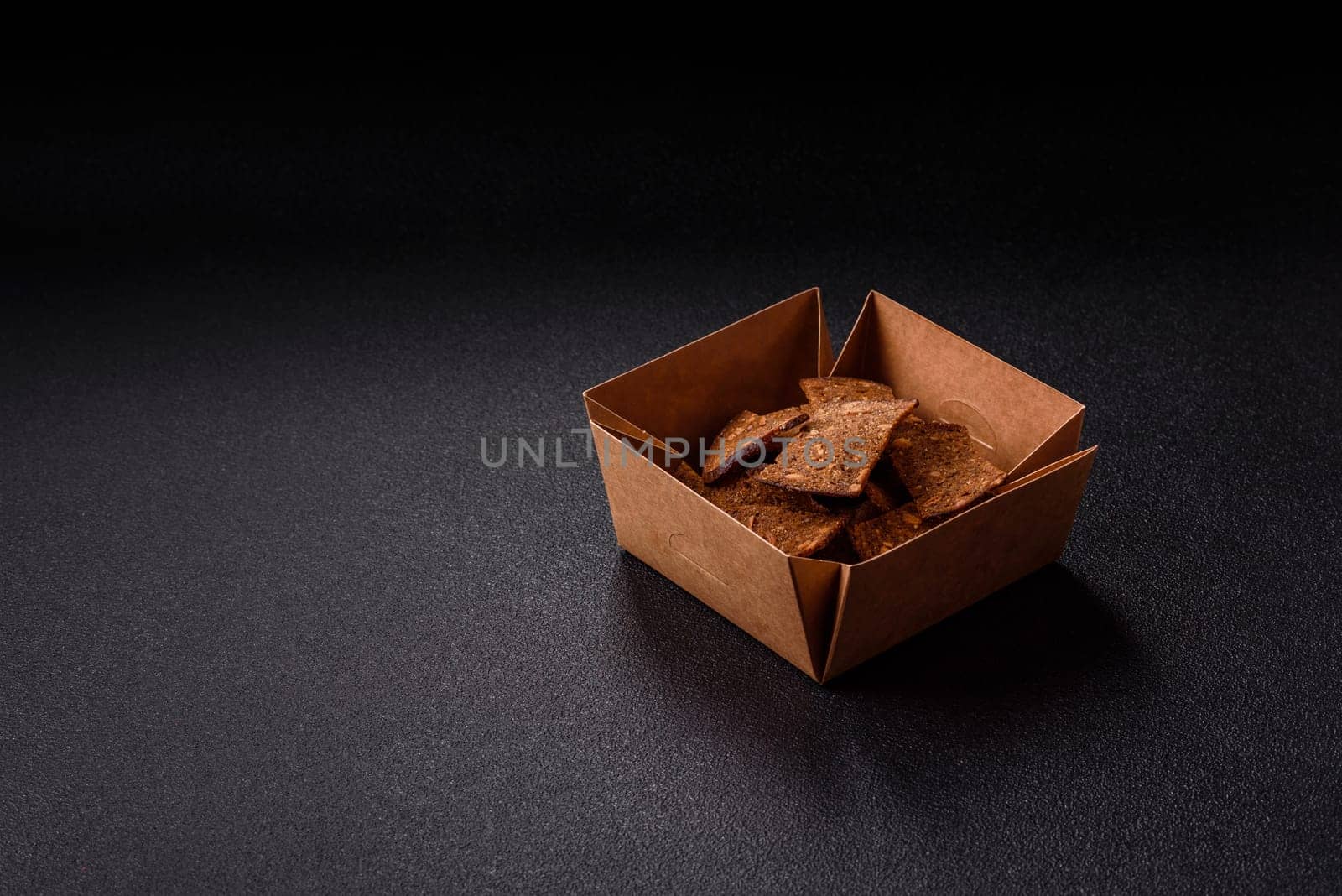 Crispy homemade brown bread chips with seeds and grains, salt, spices and herbs on a dark concrete background