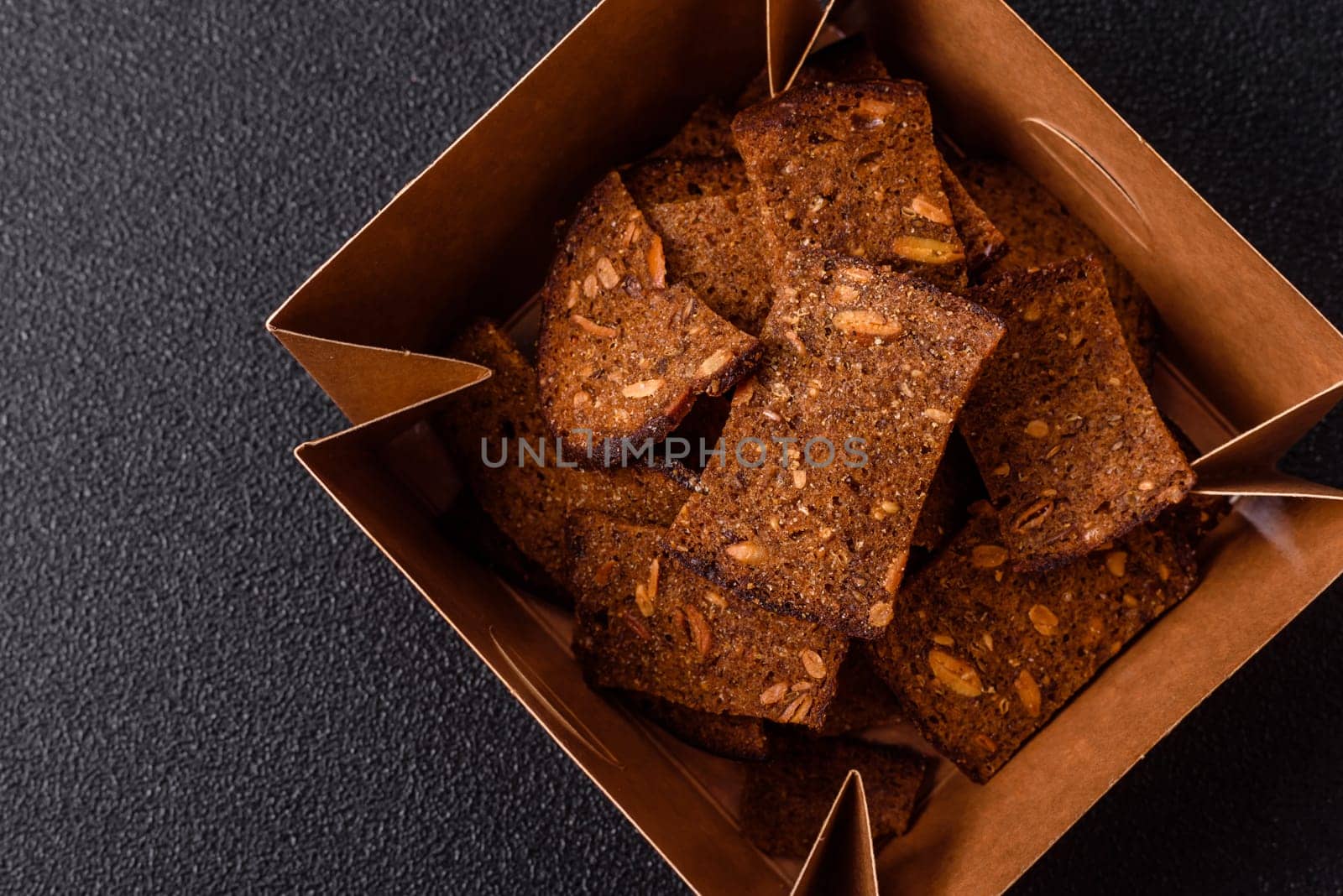 Crispy homemade brown bread chips with seeds and grains, salt, spices and herbs on a dark concrete background