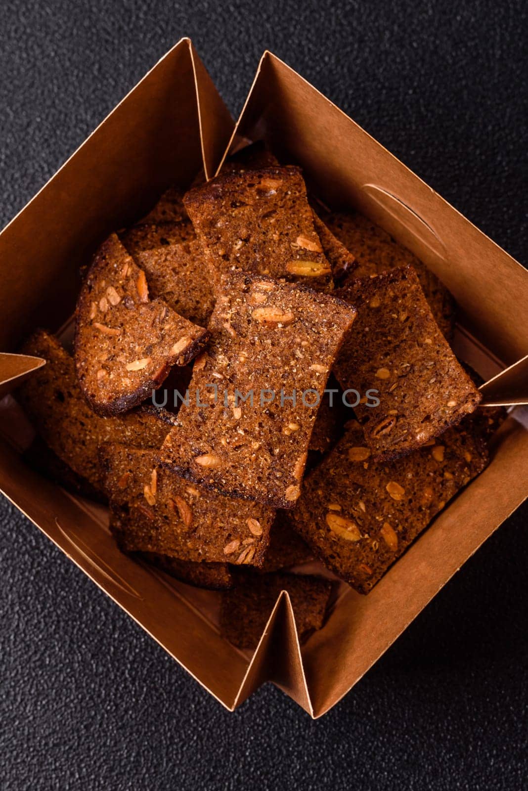 Crispy homemade brown bread chips with seeds and grains, salt, spices and herbs on a dark concrete background