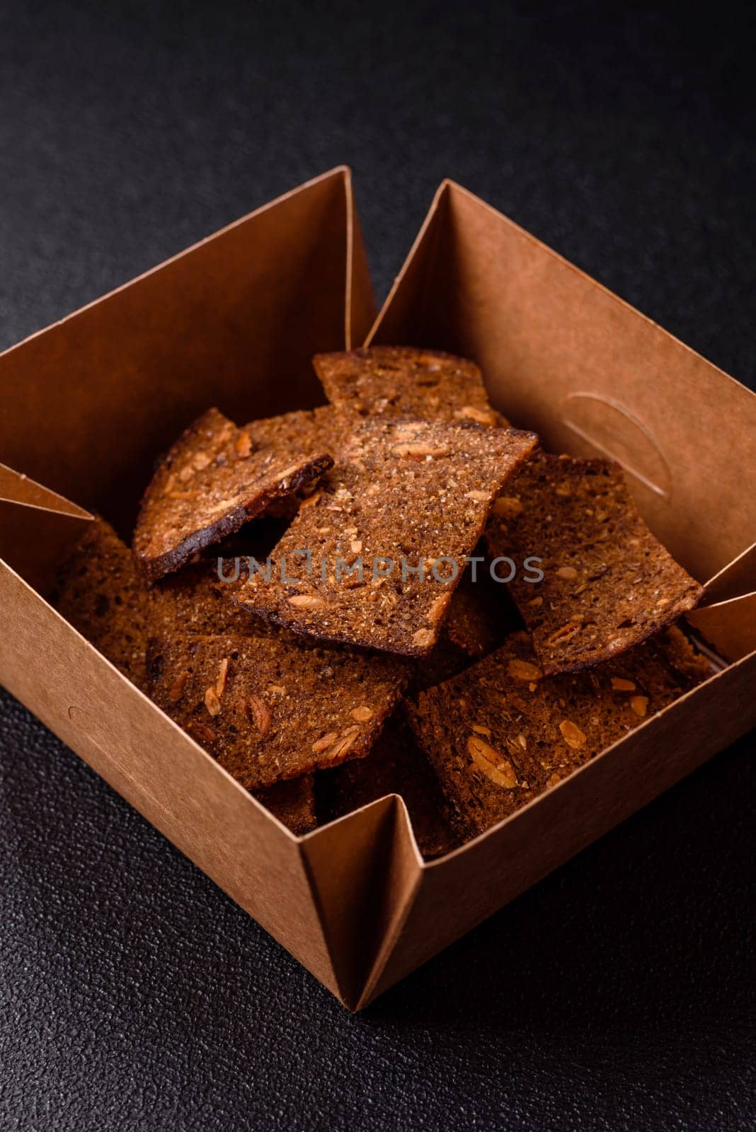 Crispy homemade brown bread chips with seeds and grains, salt, spices and herbs on a dark concrete background