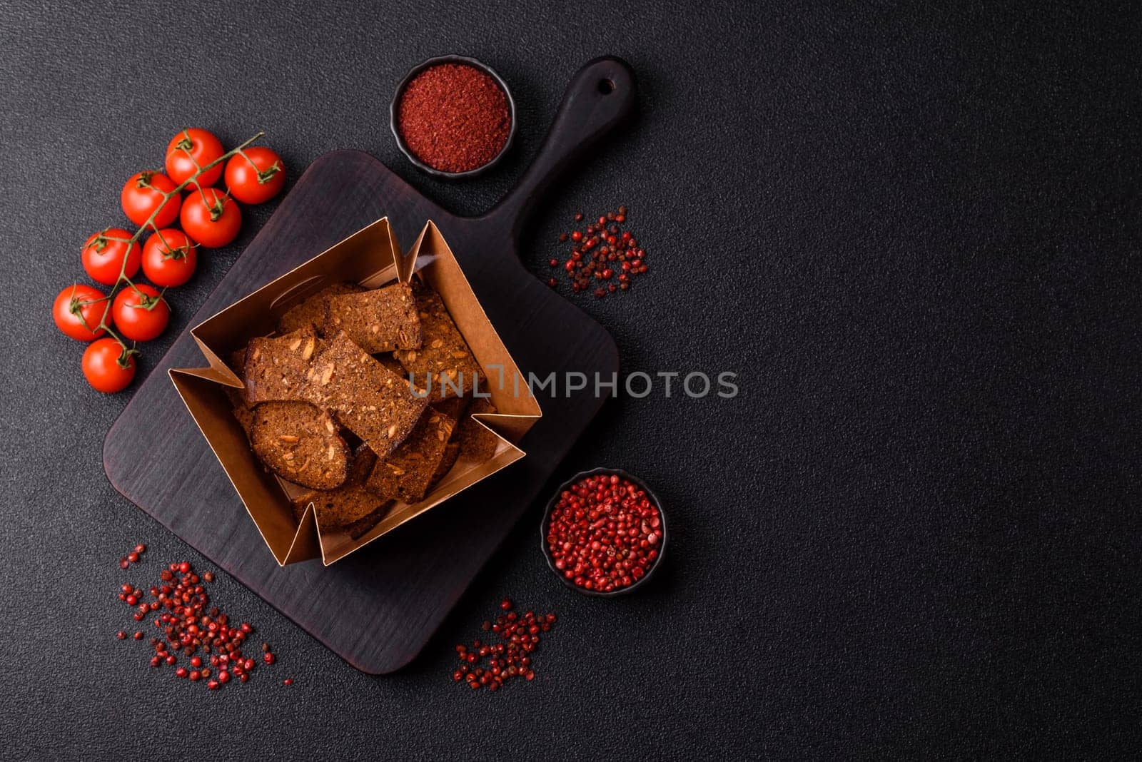 Crispy homemade brown bread chips with seeds and grains, salt, spices and herbs on a dark concrete background