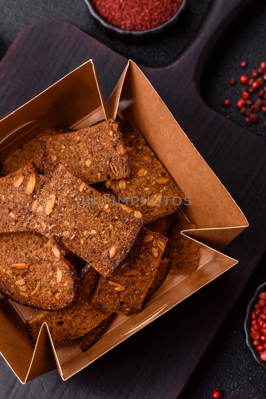 Crispy homemade brown bread chips with seeds and grains, salt, spices and herbs on a dark concrete background