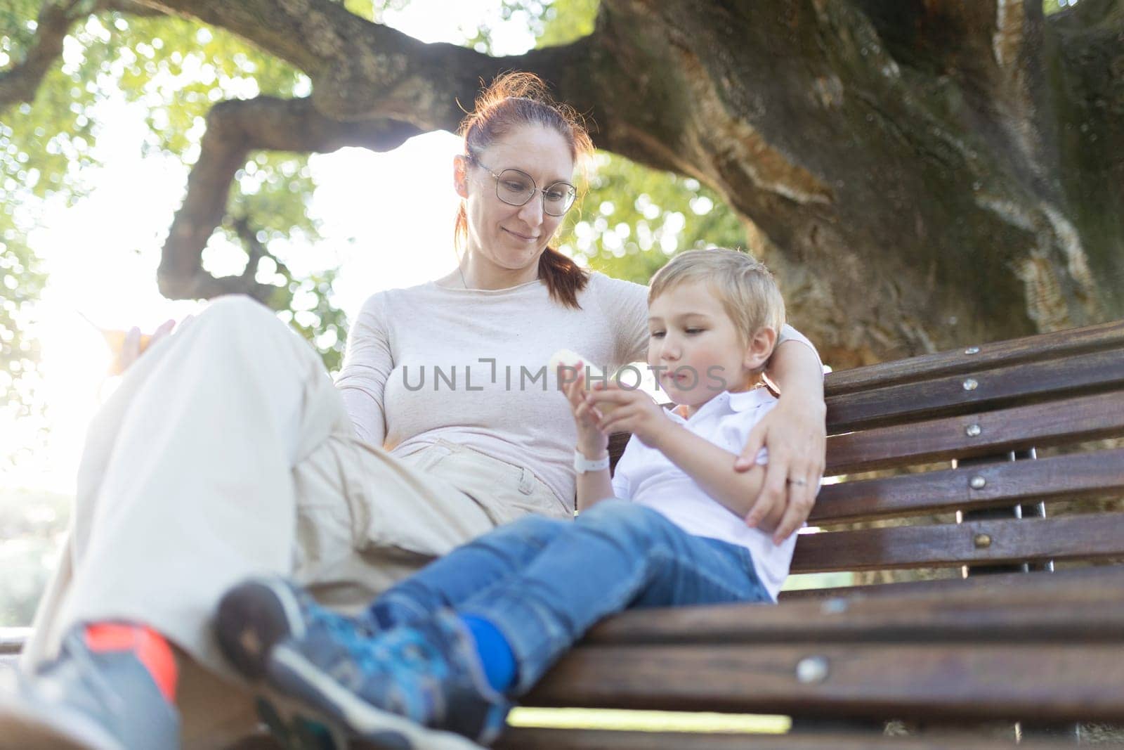 Mother and her son are enjoying a peaceful day together, sitting on a park bench under a shady tree