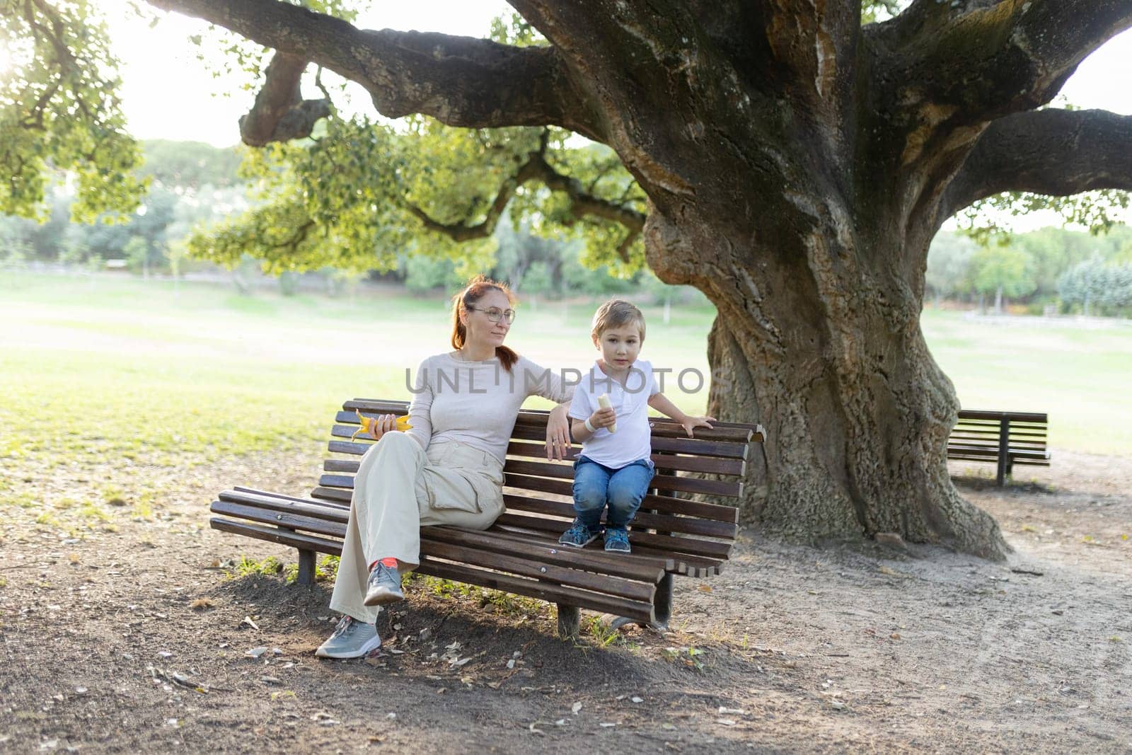 Mother and her son are enjoying each other's company while sitting on a park bench under a large tree