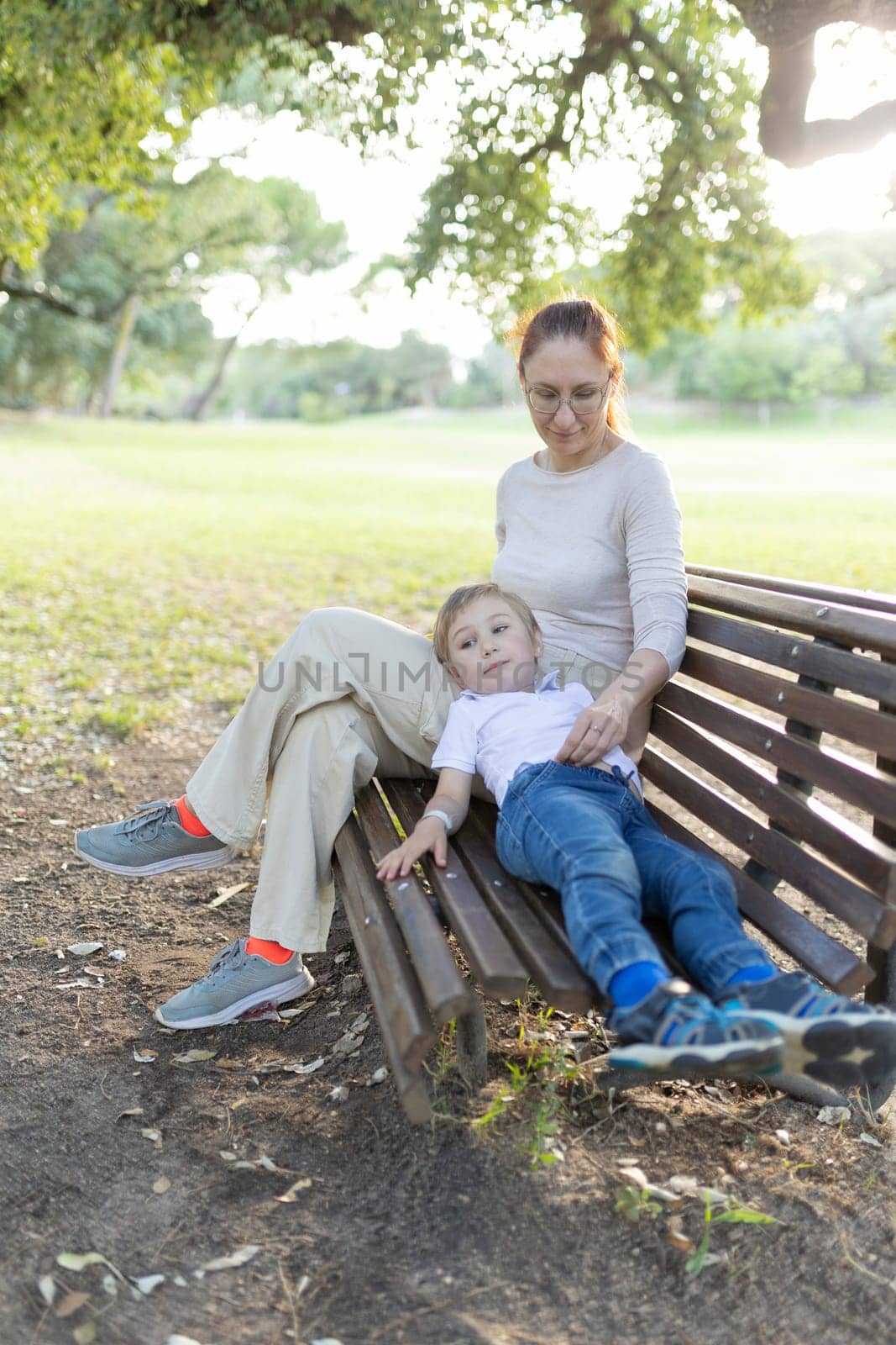Mother sits on a park bench on a summer day while her son lays across her lap
