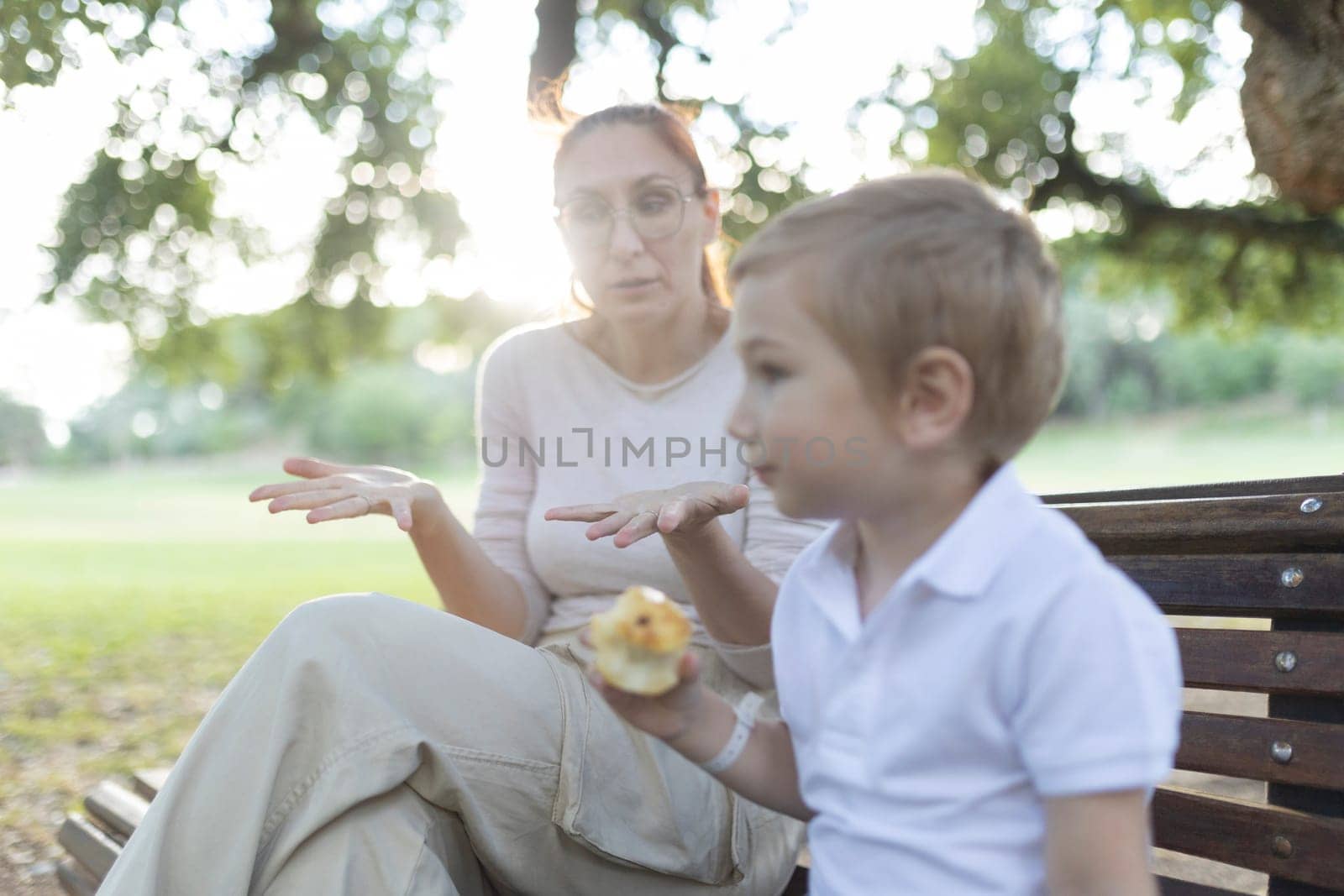 Mother is gesturing with her hands while talking to her son, who is holding a snack, in a park