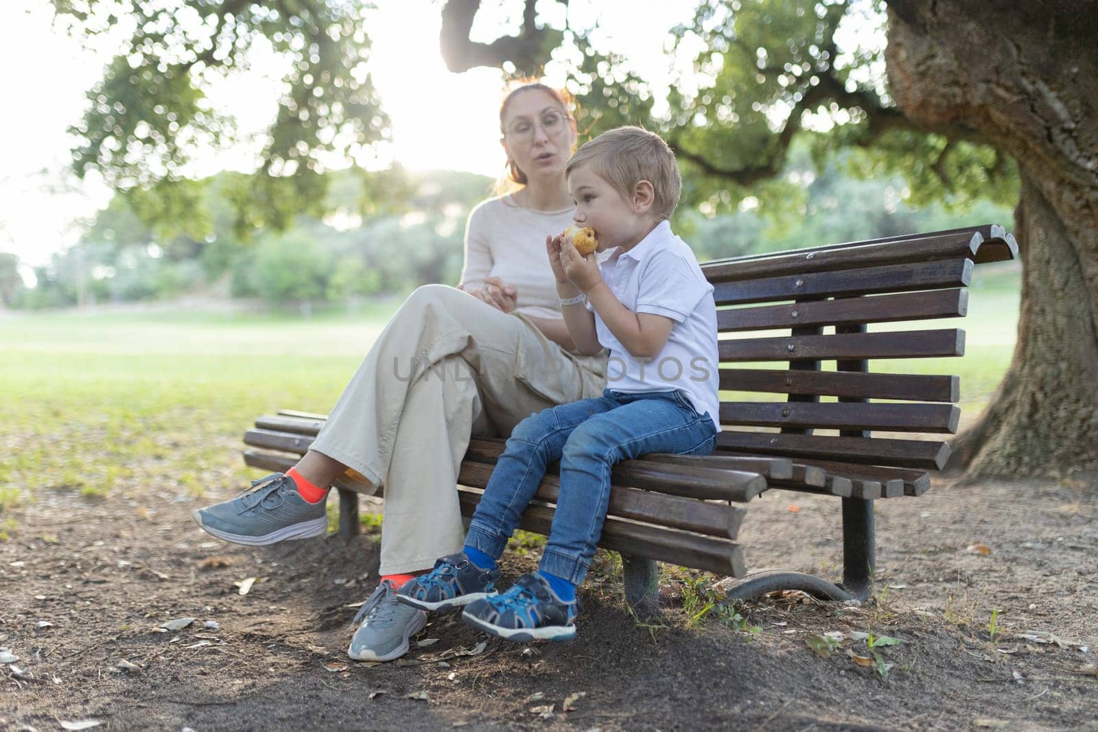 Mother is talking to her son while he eats a snack on a park bench