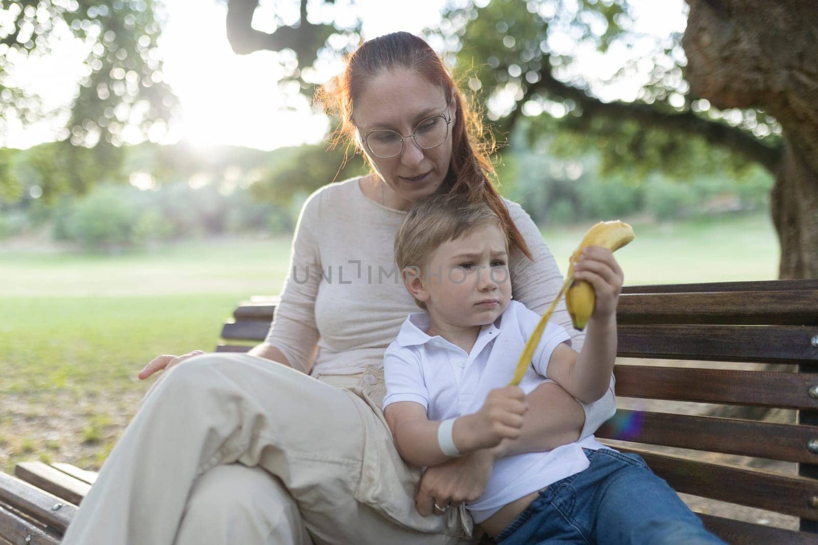 Young child is holding a banana while talking with his mother on a park bench in the summer