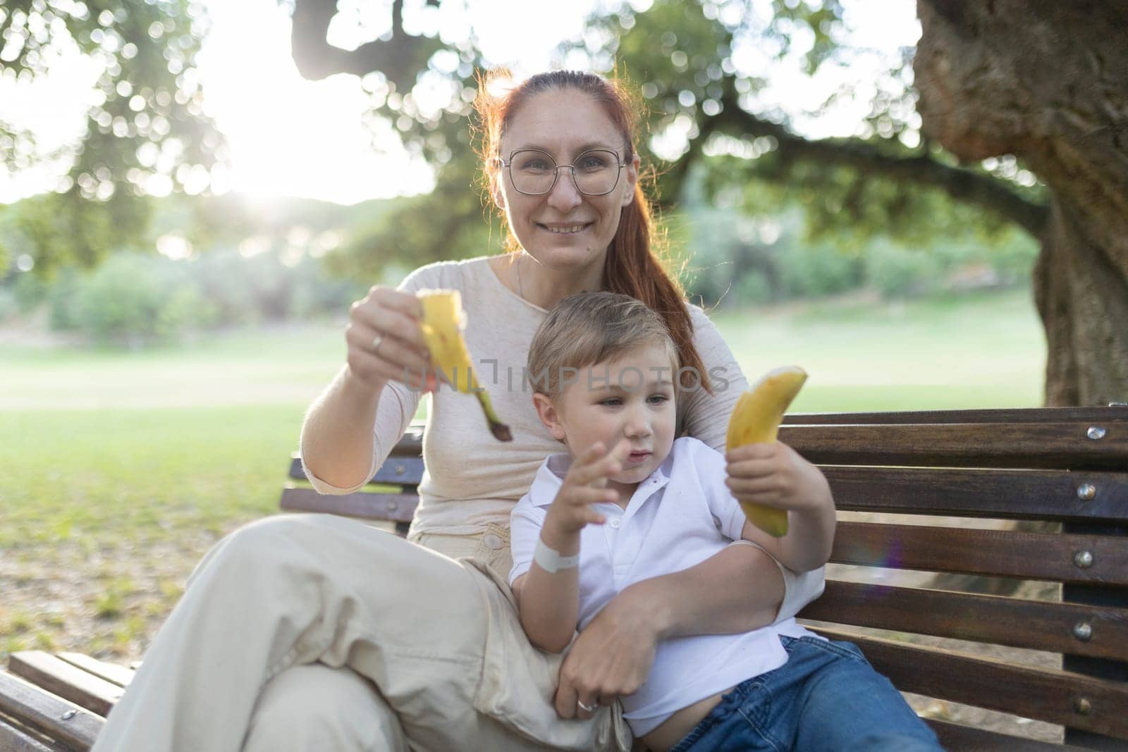 Mother and her son are sitting on a park bench, eating bananas and enjoying a sunny day together