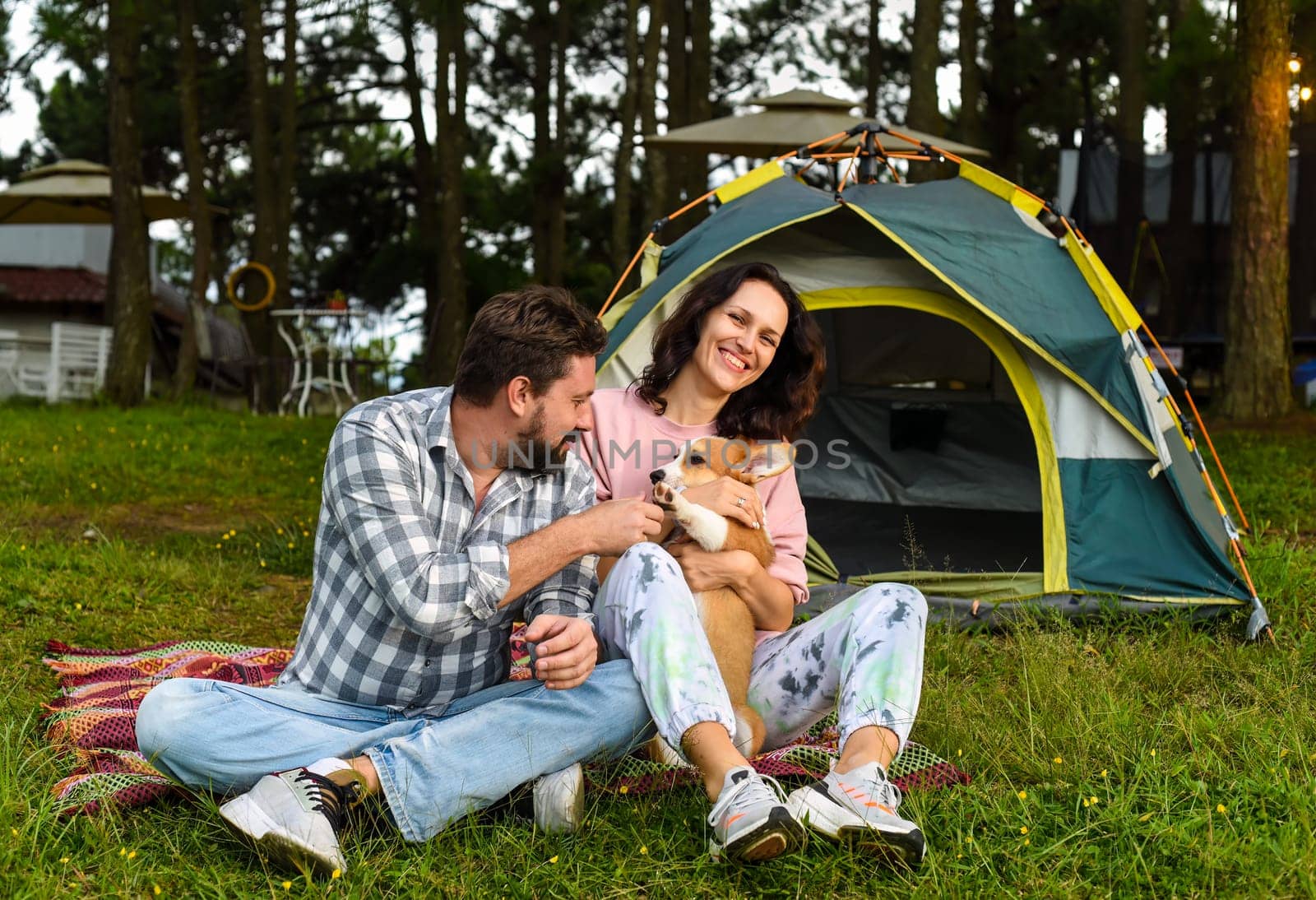 Young married couple with their corgi dog sitting in the forest near tent and looking at sunsetYoung married couple with their corgi dog sitting in the forest near tent and looking at sunset