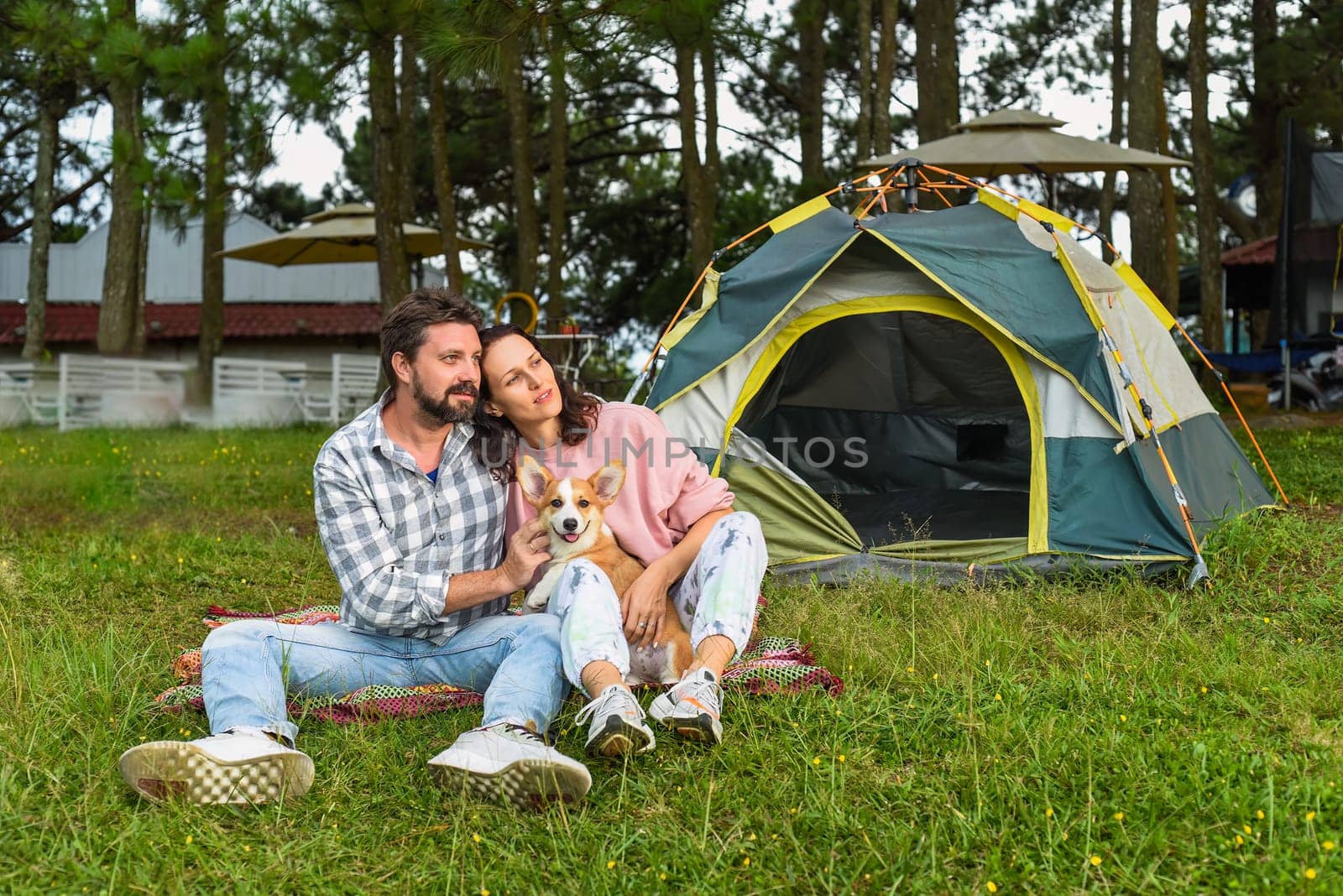 Young married couple with their corgi dog sitting in the forest near tent and looking at sunset