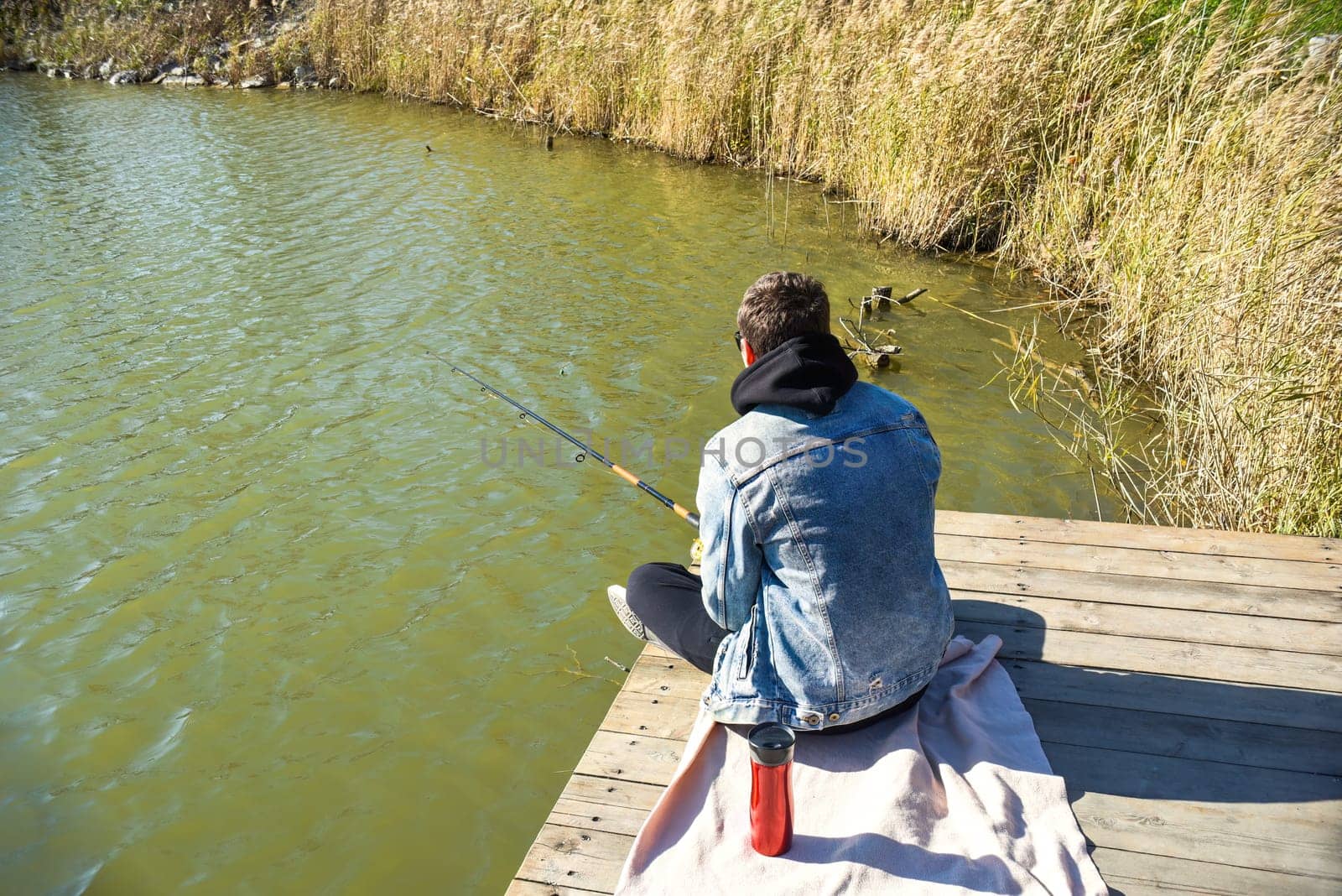 Young fisherman in denim jacket sitting on wooden pier, fishing in the lake. he holding the rod and looking at float.