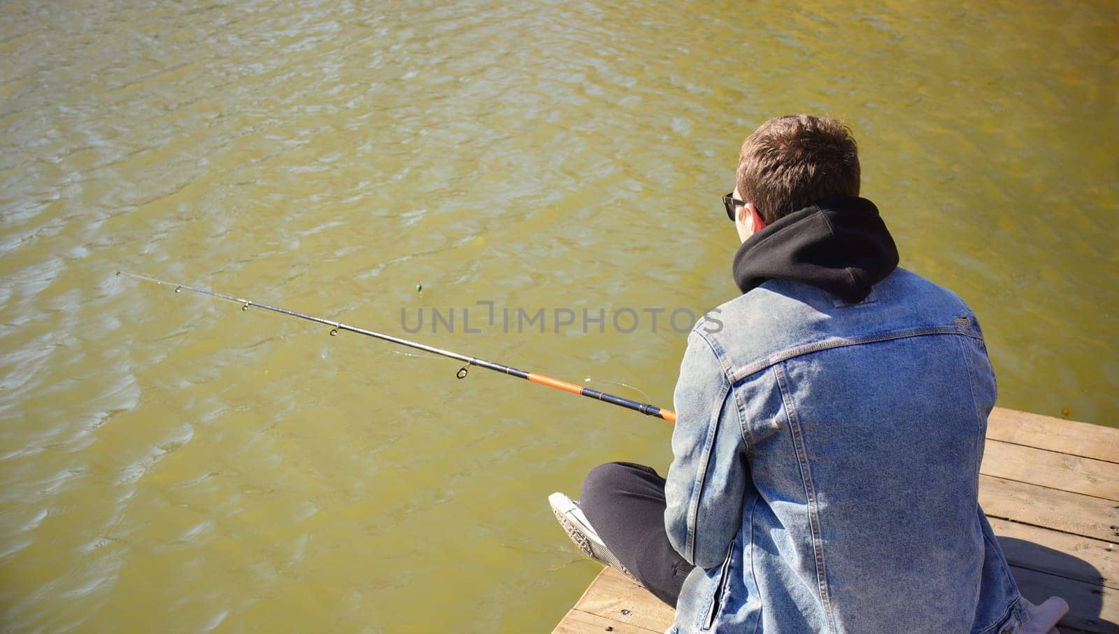 Young fisherman in denim jacket sitting on wooden pier, fishing in the lake. he holding the rod and looking at float.