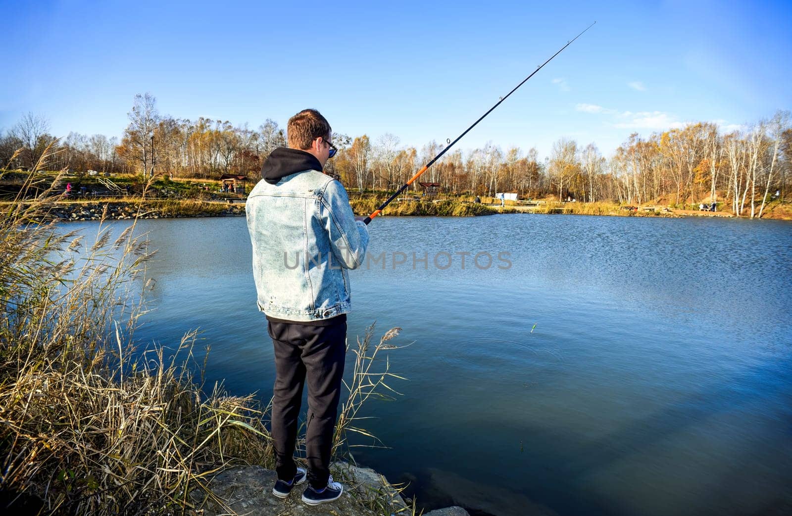 Young fisherman in denim jacket standing on stone, fishing in the lake. he holding the rod and looking at float against autumn forest in park.