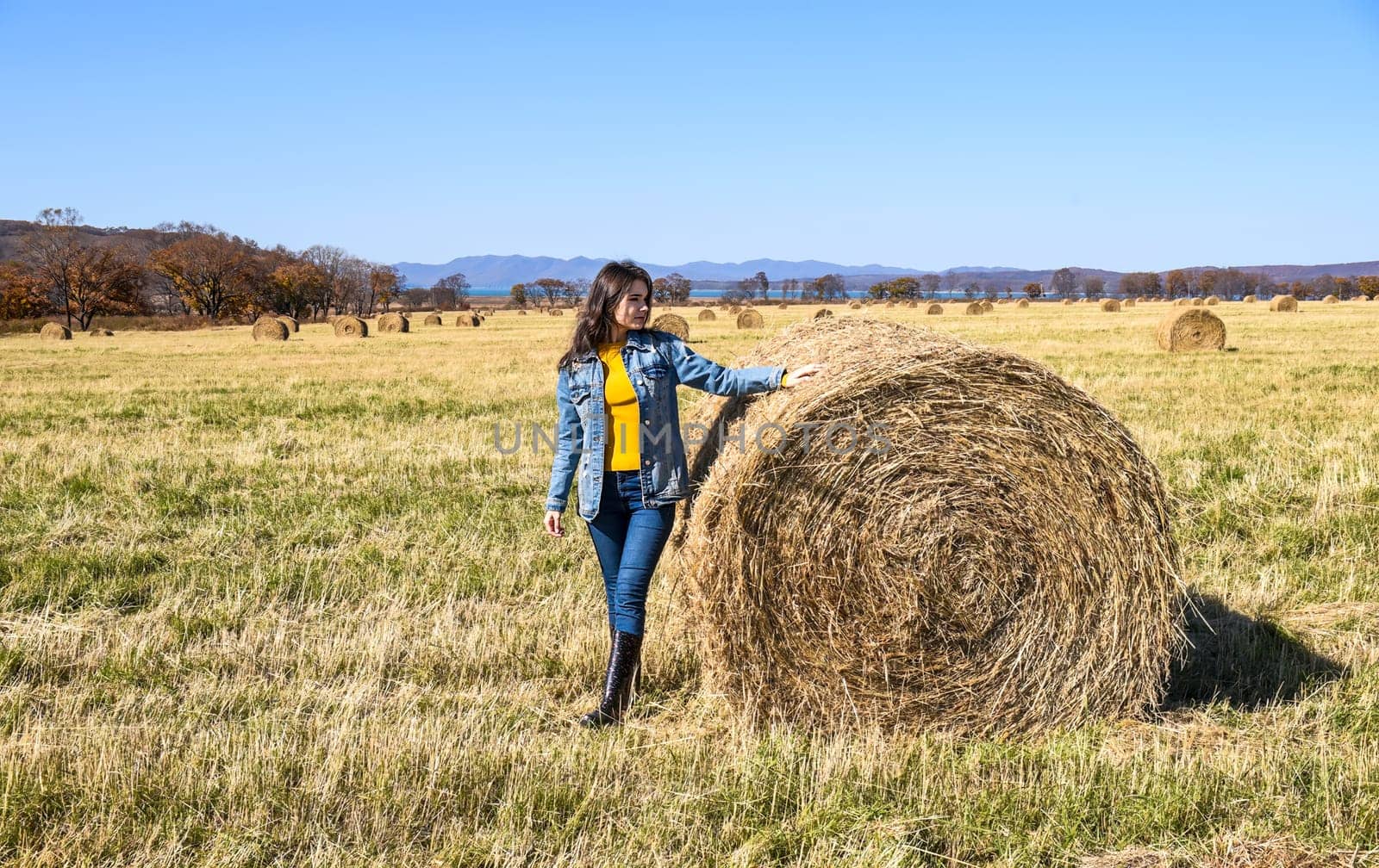 Young brunette woman in denim jacket standing near haystack in field with bales of hay