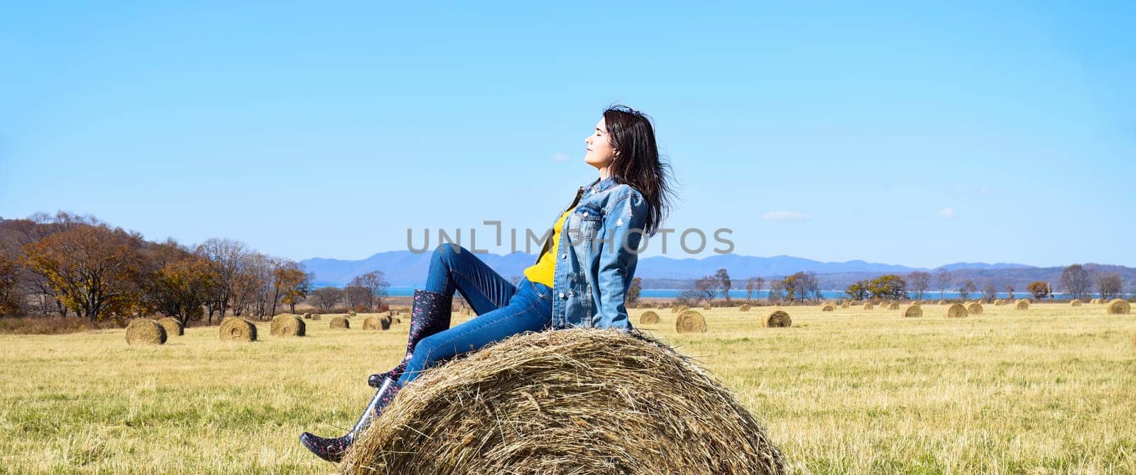 Young brunette woman sitting on top of haystack in field. she squinting from the sun