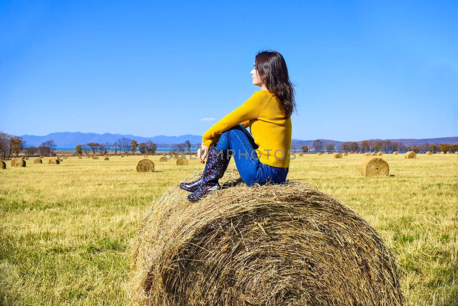 Young brunette woman in yellow sweater and rubber boots sitting on top of haystack in field. she looking at mountains, Japanese sea and the autumn forest. Primorsky kray, Russia.
