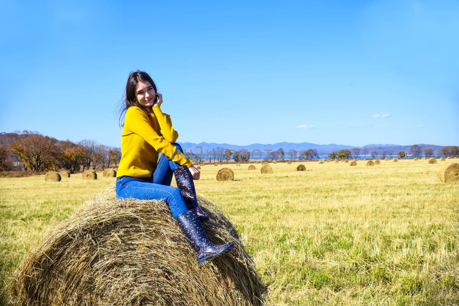 Young brunette woman in yellow sweater and rubber boots sitting on top of haystack in field