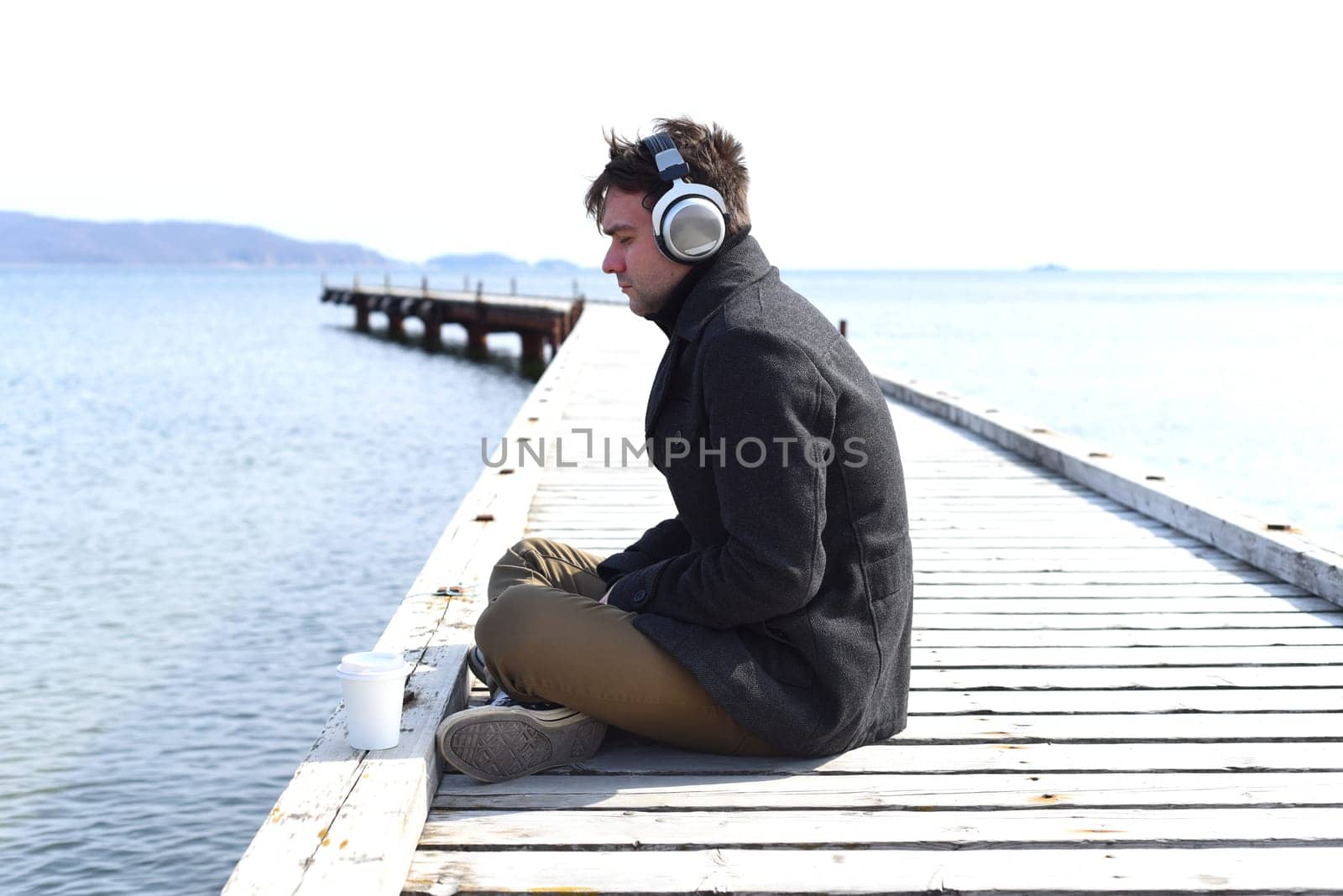 Young gloomy man listening to music sitting all alone on pier on a windy spring day.