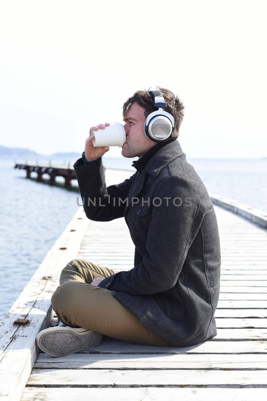 Young man listening to music and drinking coffee, sitting alone on pier on a sunny day