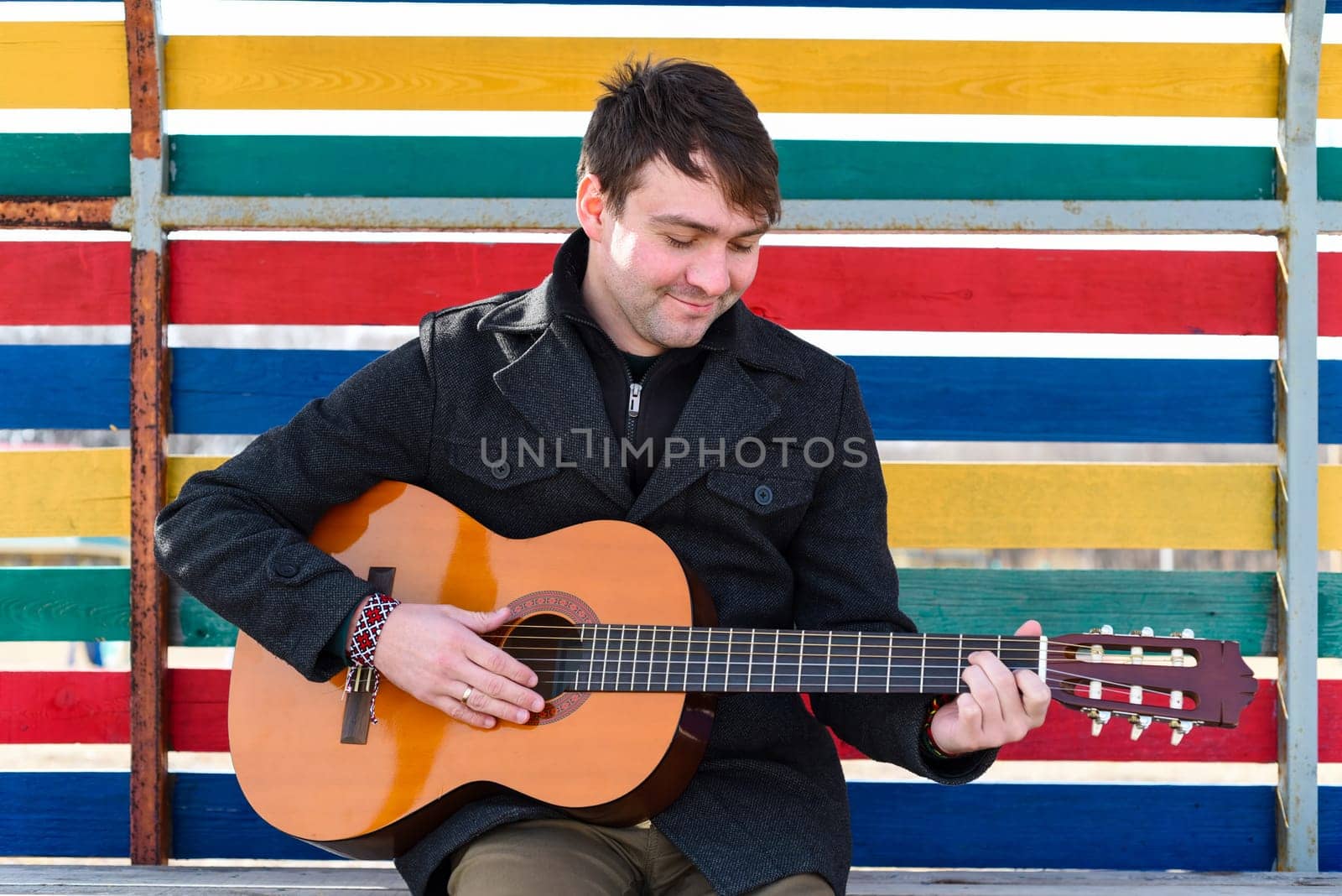 Smiling young man playing guitar and sitting against colorful wall