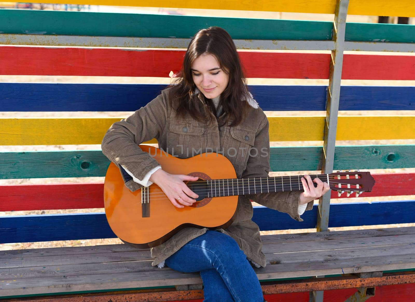 Young woman playing guitar and laughing, sitting on a bench against colorful background