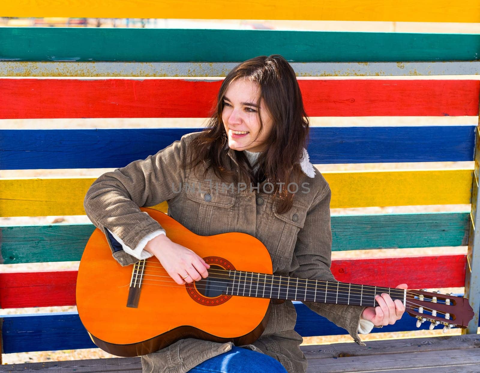 Young woman playing guitar and laughing, sitting on a bench against colorful background