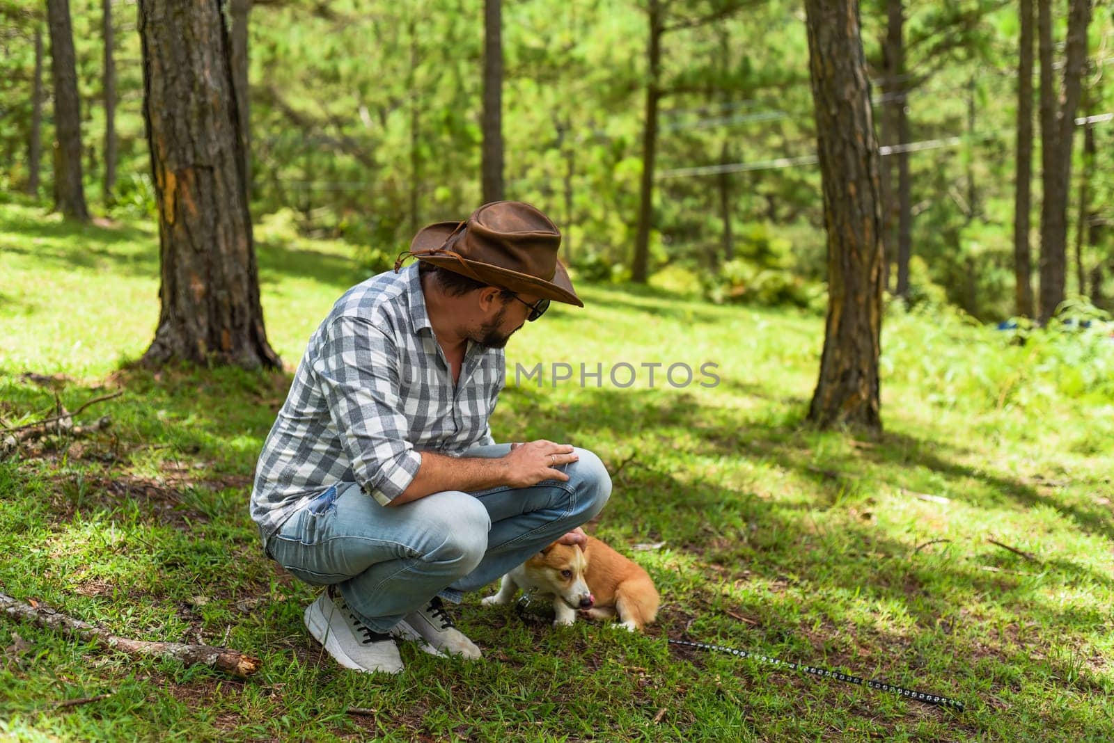 Young man in hat touching and playing with his corgi dog in forest