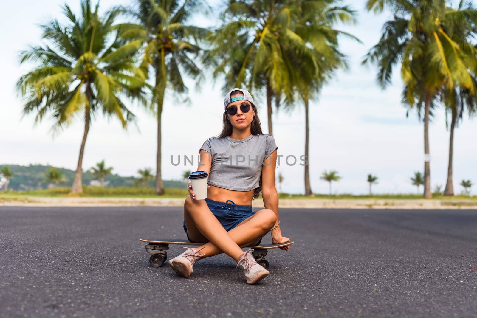 Young beautiful woman in a cap sitting on a skateboard and drinking coffee