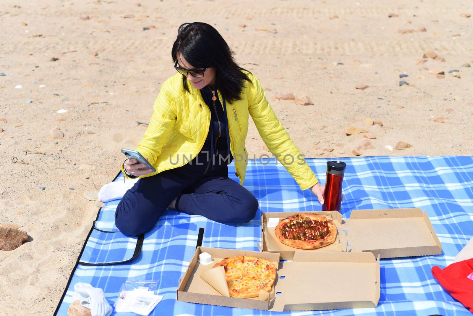 Russian 45 year old woman going to eat pizza while scrolling on phone, sitting on the beach