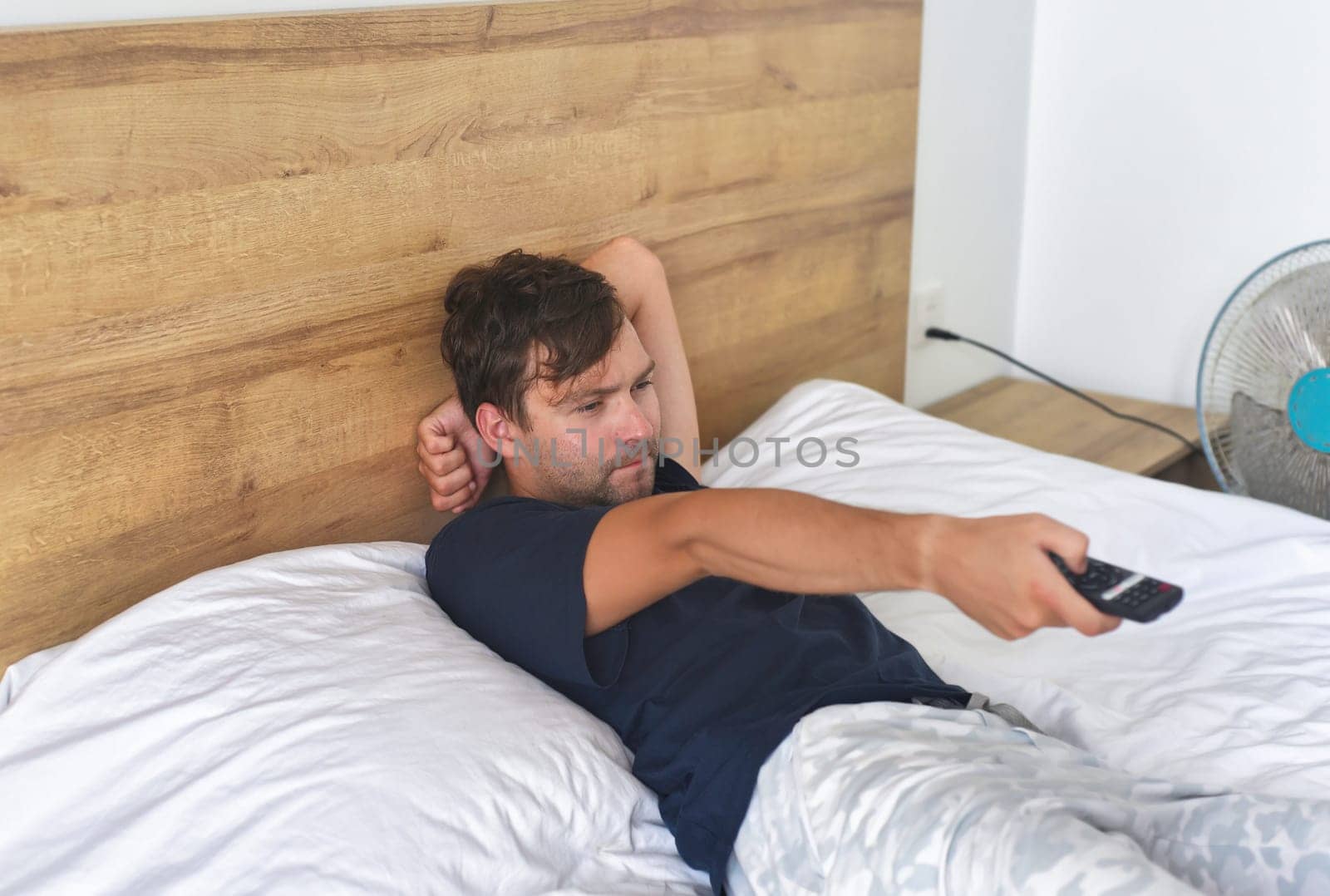 Man watching TV  and holding a TV remote on a bed with fan