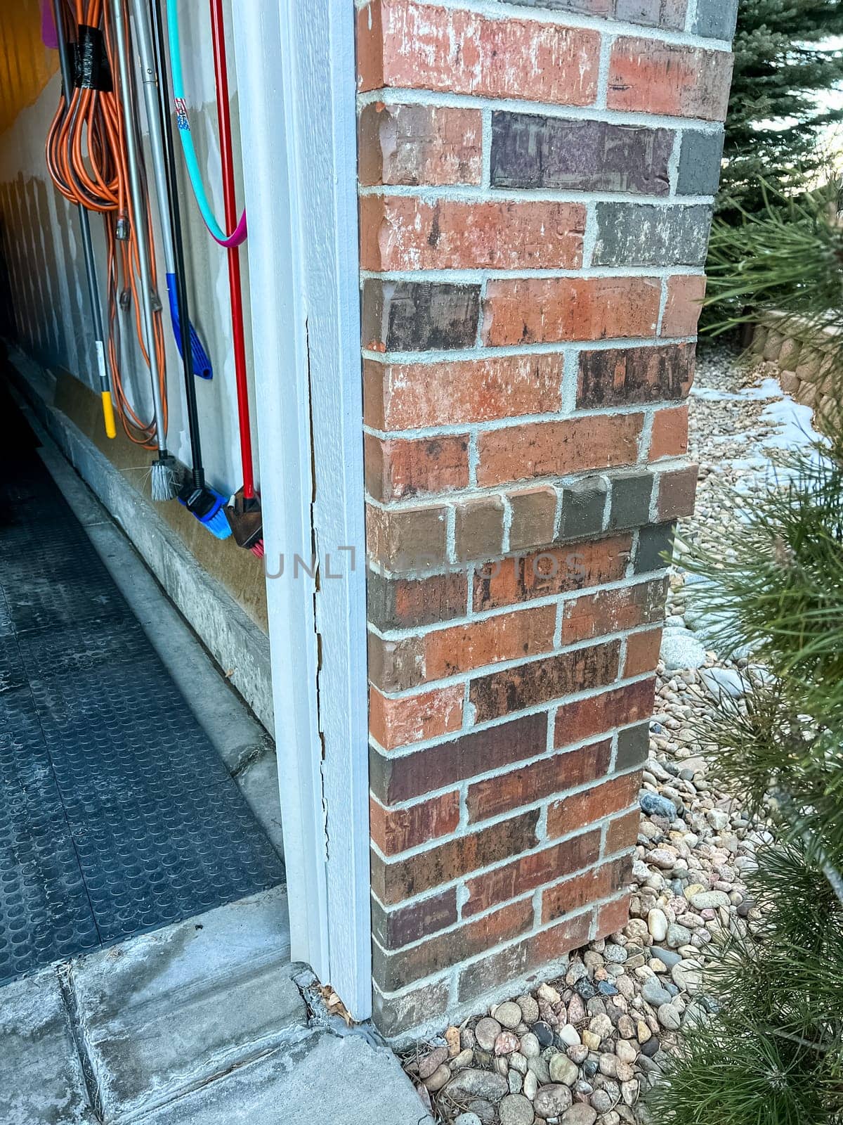 Castle Pines, Colorado USA - January 10, 2023-A close-up view of a garage entrance shows a cracked white trim running vertically along the brick corner The damage is visible near the base where the trim has separated slightly from the structure Inside the garage various tools and extension cords are hanging on the wall The ground is covered with rubber matting while the exterior has decorative rocks and a pine tree nearby