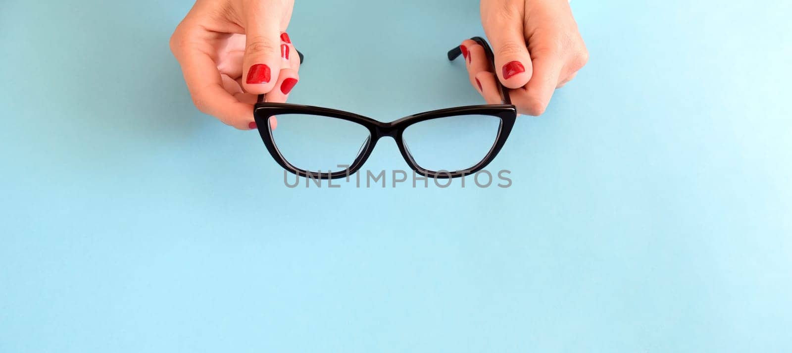 Female hands with red nail polish holding black glasses for eye vision with frame Cat's eye on a blue background. Copy space