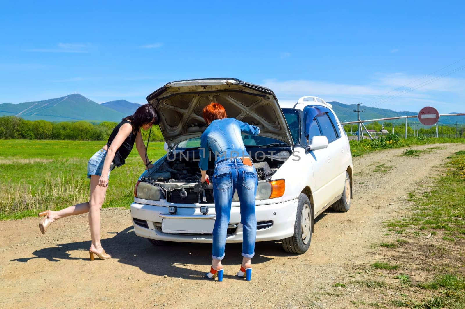 Red-haired woman with a screwdriver, on high heels standing near a broken car with open hood and brunette girl on a country road and barrier with stop sign