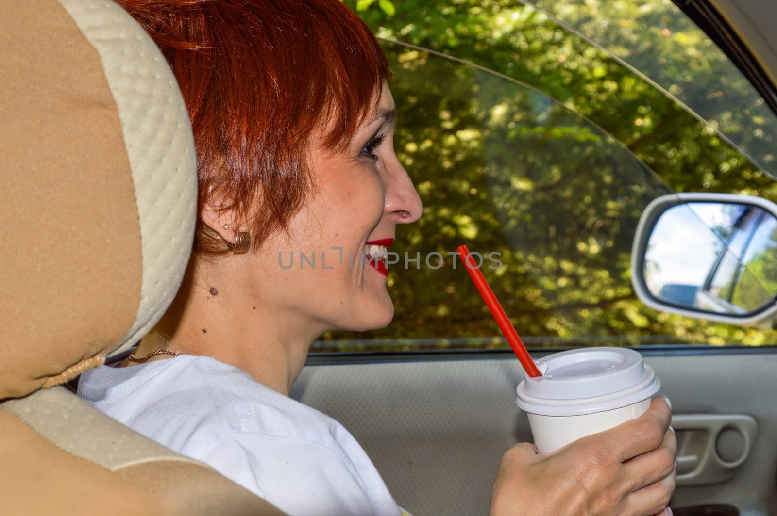 Cheerful young woman passenger with red hair sitting in the car, holding plastic cup of coffee and smiling with teeth. Summer adventures