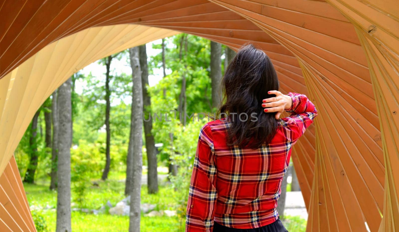 Brunette woman in red shirt standing with her back to the camera and holding her hair in hand inside the wooden tunnel with arches. path to the forest