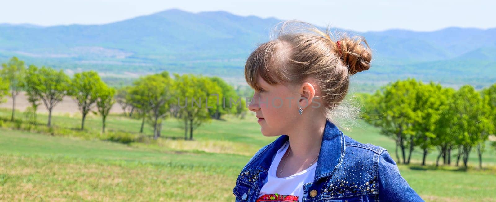 Portrait of 10 year old Russian smiling blonde girl with hairstyle bun on a background of green field with trees. cute face, looking left