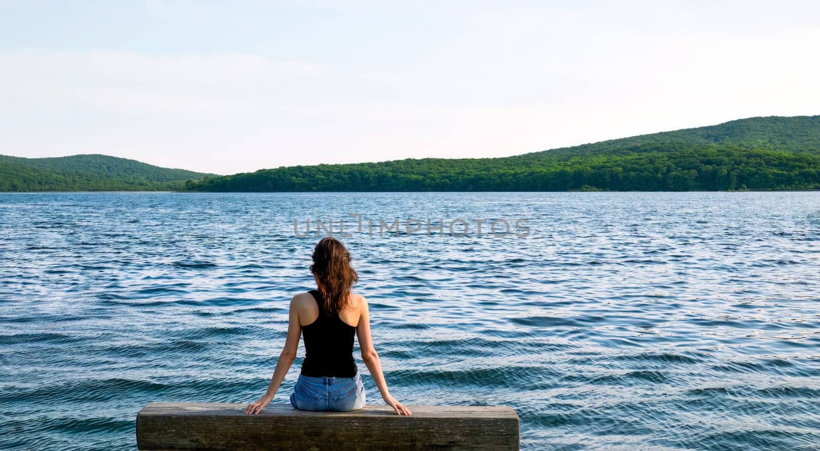 Brunette woman sitting on wooden bench on the pier on a dark evening  against the background of the sea and forest. Copy space on top and side. Novik Bay, Sea of ​​Japan