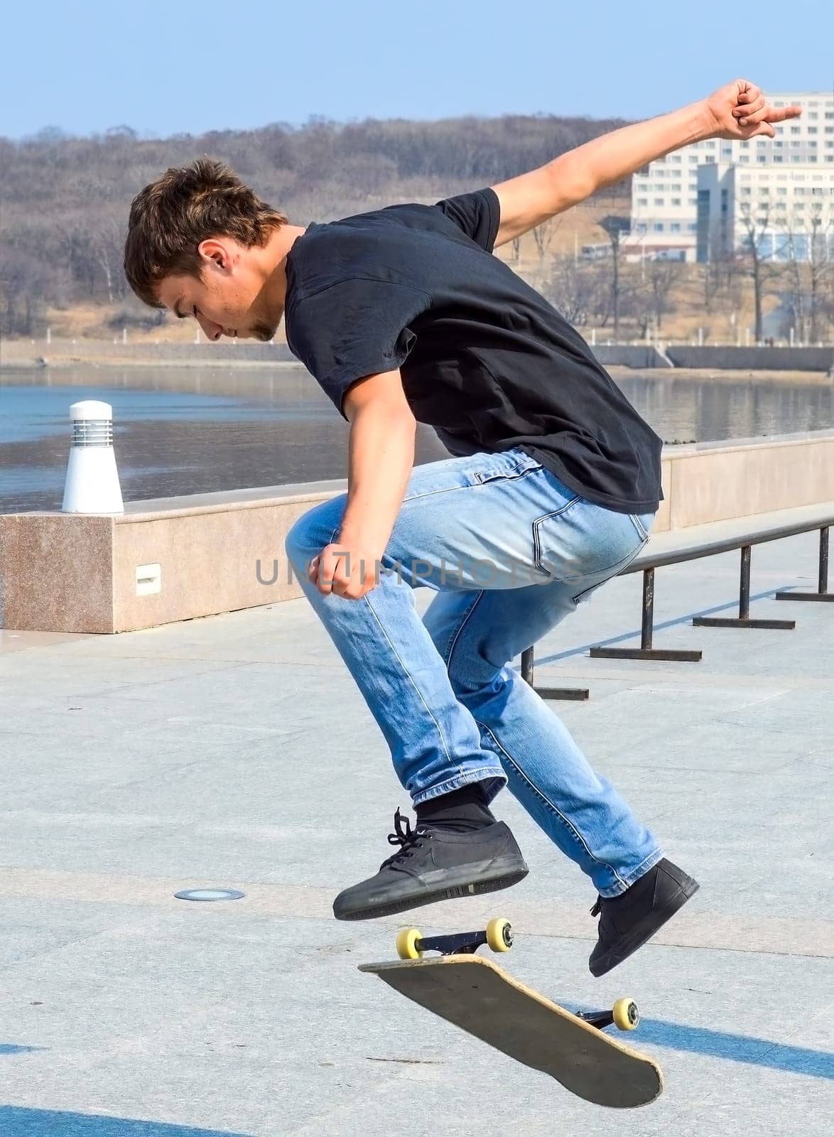 Young Russian skater in black t-shirt and blue jeans jumping on the skateboard on a early spring day